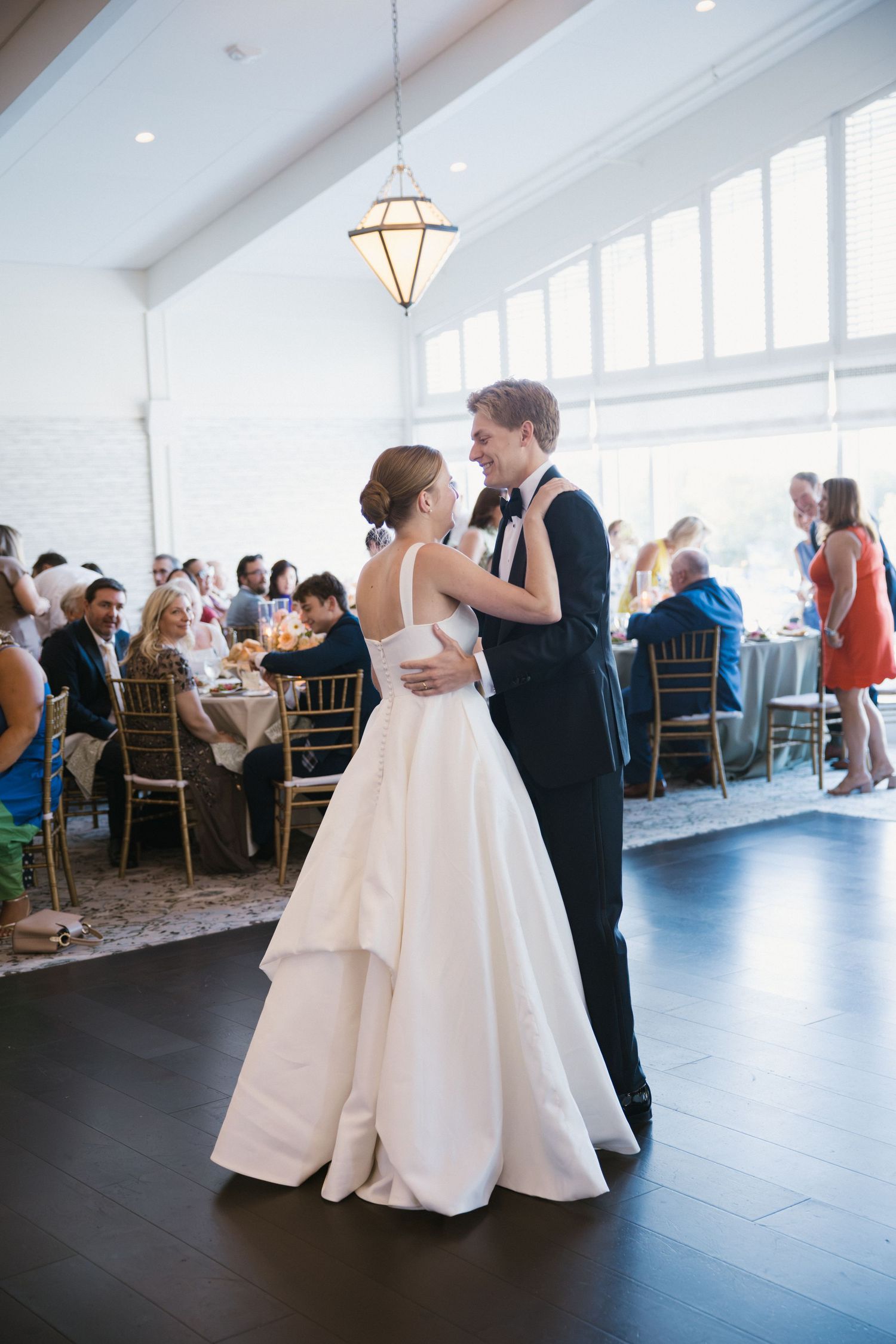 Bride and groom share first dance in elegant white reception hall surrounded by seated guests.