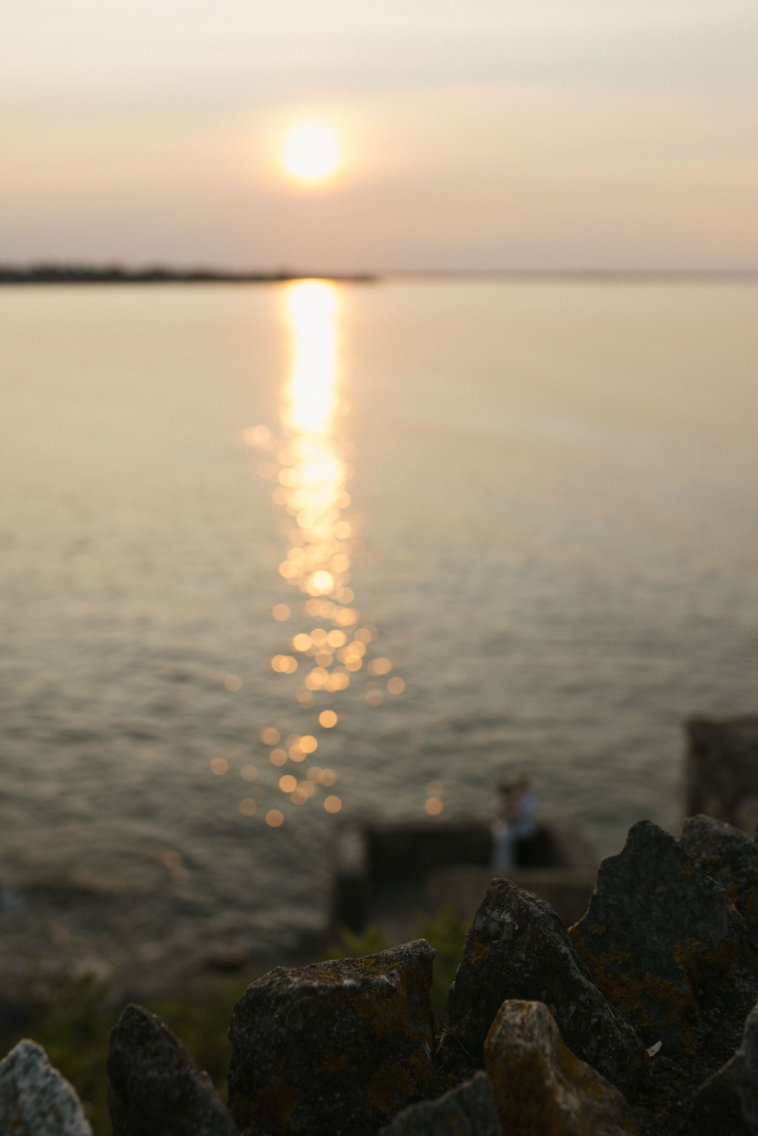 Blurred rocks in foreground with golden sunset reflecting on calm ocean waters at dusk.