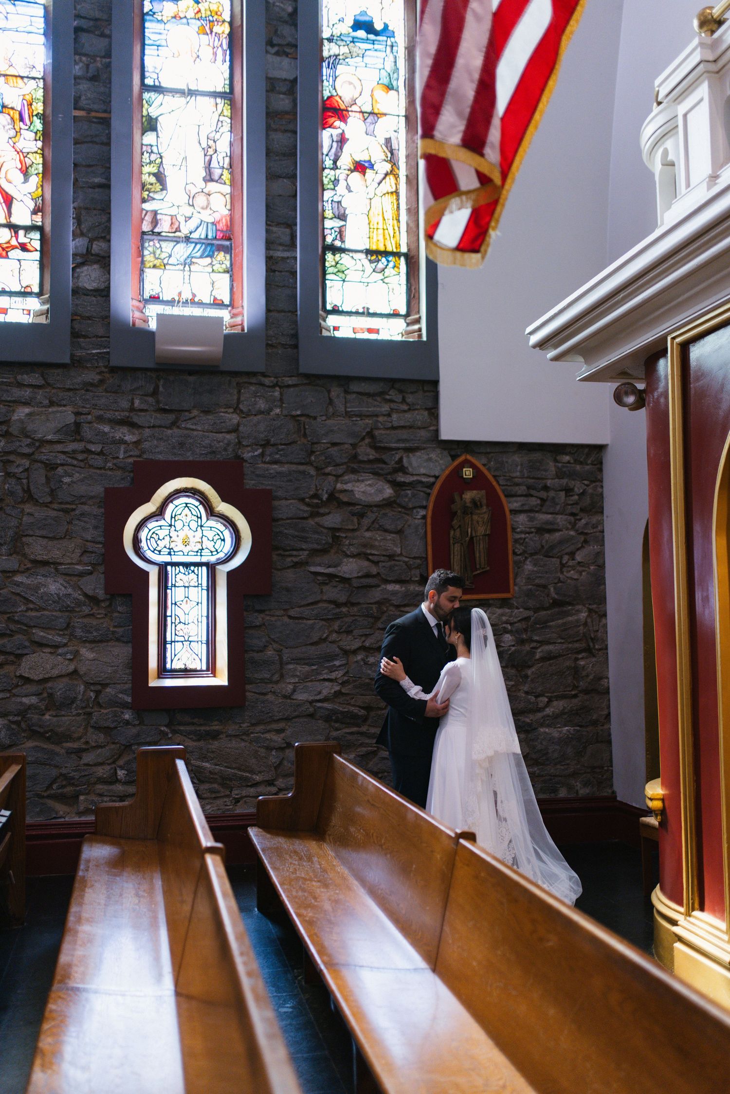 Bride and groom embracing inside stone church with colorful stained glass windows.