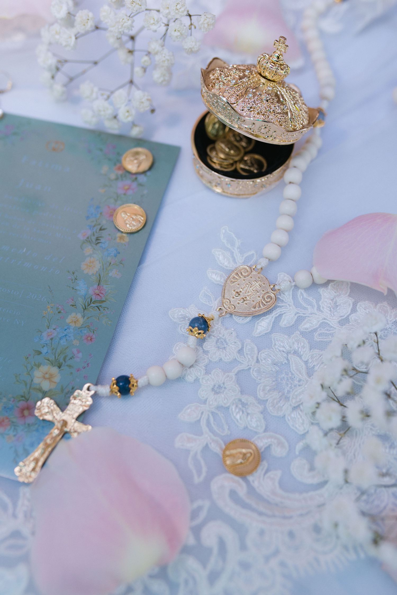 Gold rosary beads with crucifix, ornate jewelry box, coins, floral book, and rose petals on lace fabric.