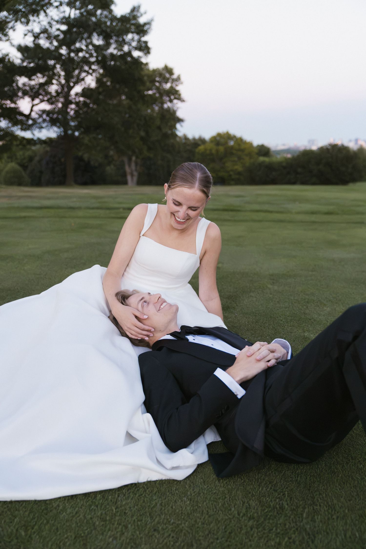 Smiling bride cradles grooms head in her lap while he lies on a green lawn outdoors.