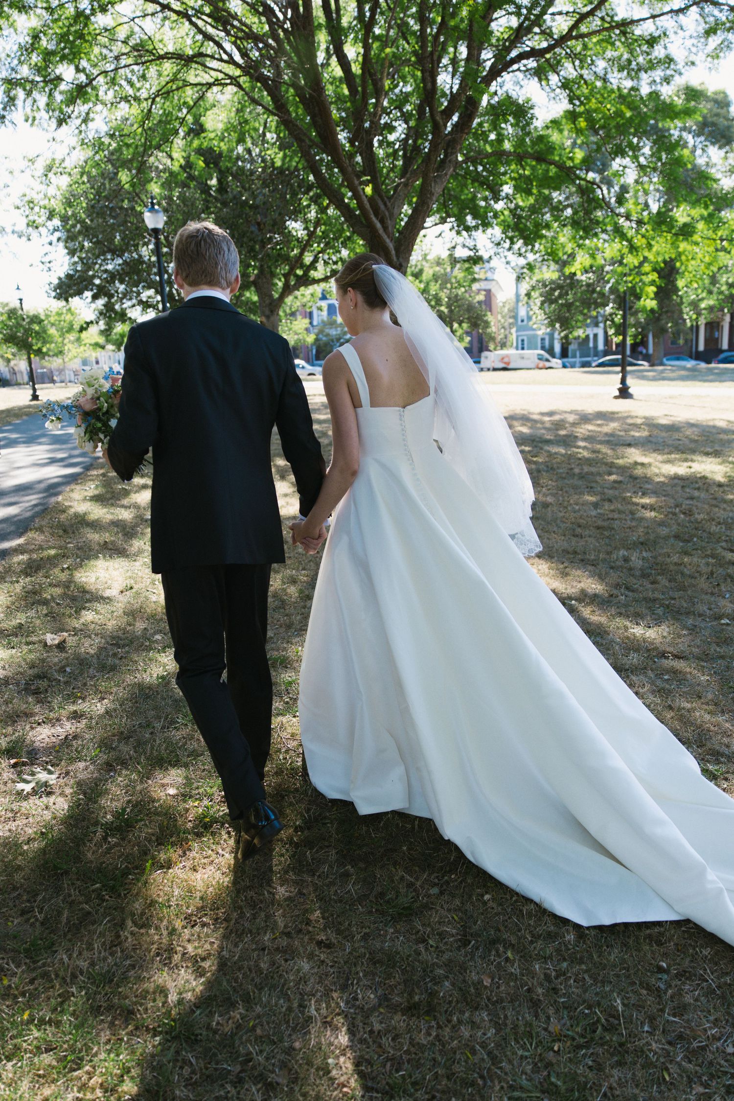 Bride and groom walk hand-in-hand through a tree-lined park, bride wearing a flowing white gown with long train.