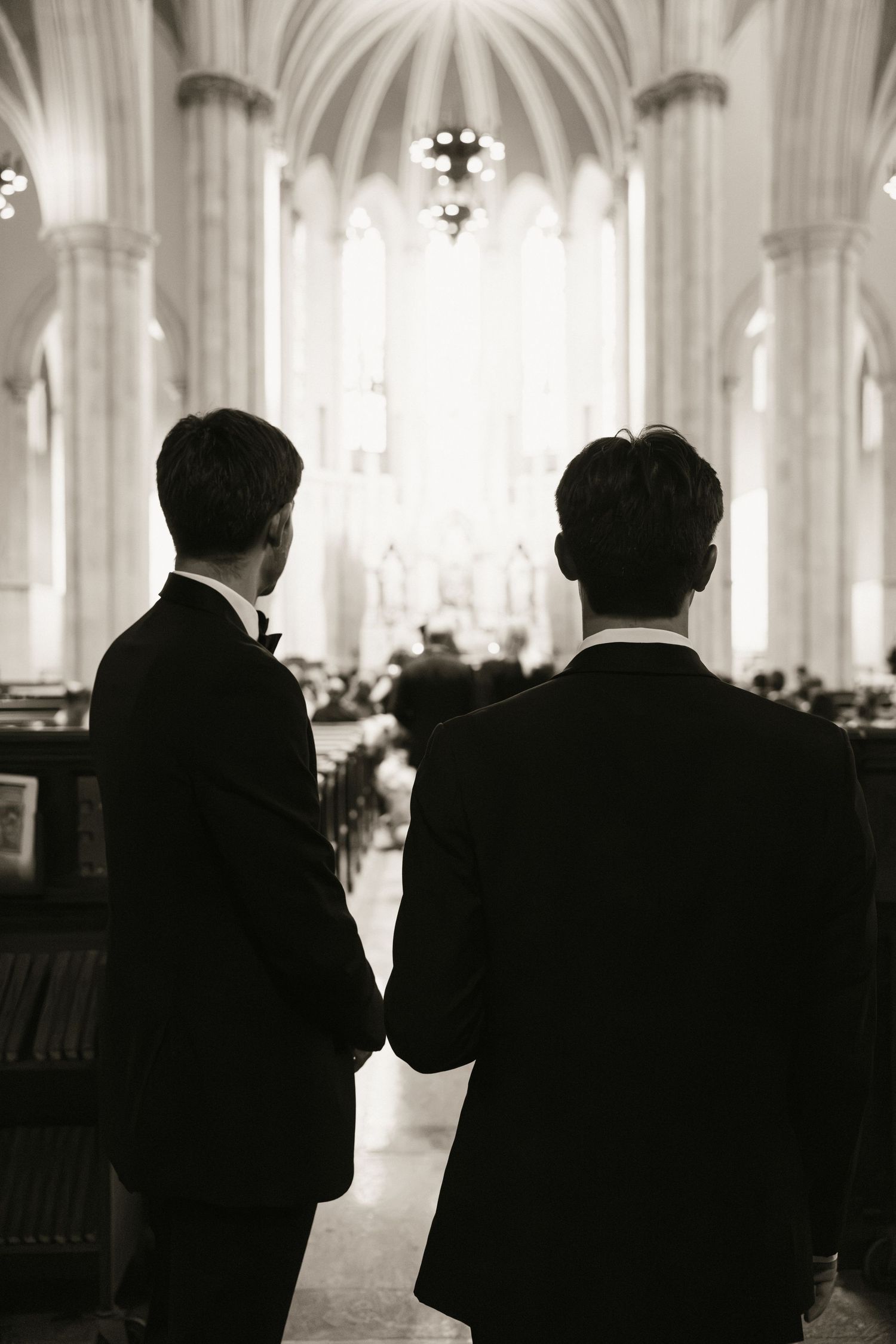 Two grooms in black tuxedos hold hands at the altar of an elegant church in a black and white wedding photo.