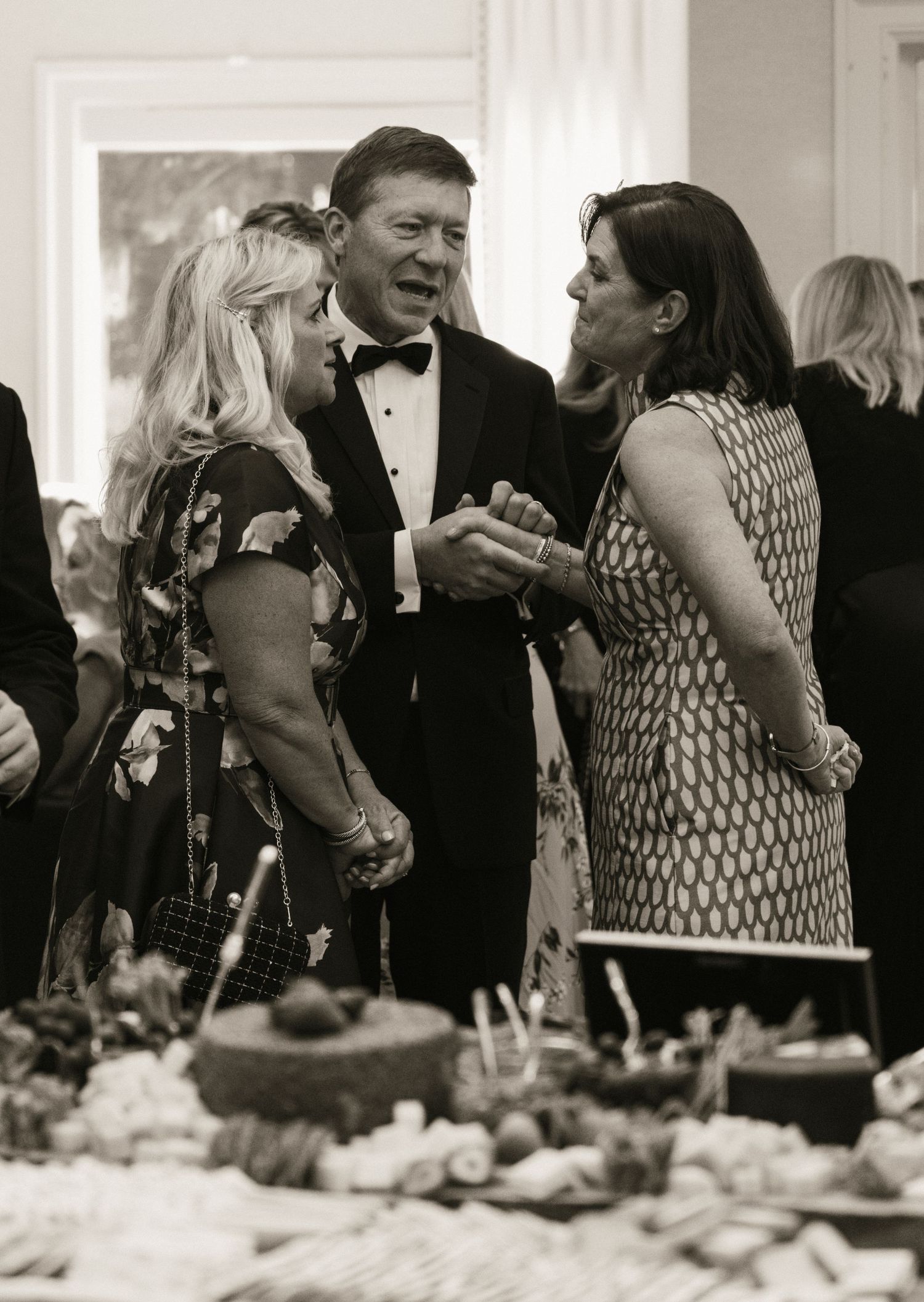 Black and white photo of wedding guests in formal attire conversing at elegant reception event.