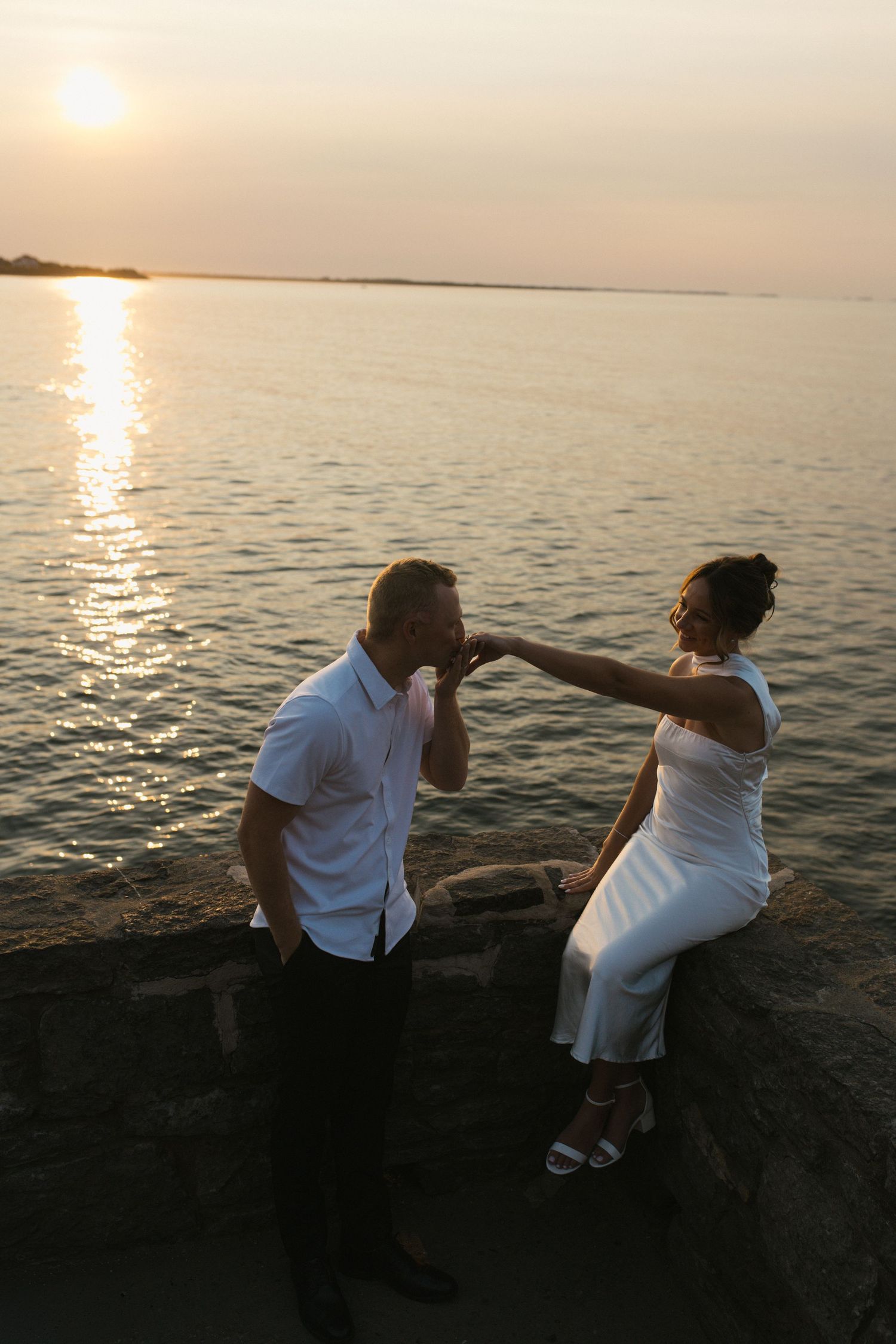 Romantic couple at sunset by the sea, man kissing womans hand while she sits on stone wall in white dress.