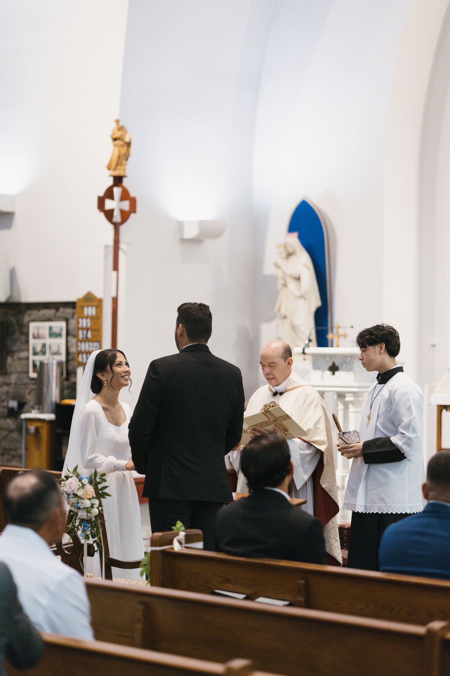 Smiling bride and groom exchange vows before priest and altar server during Catholic church ceremony.