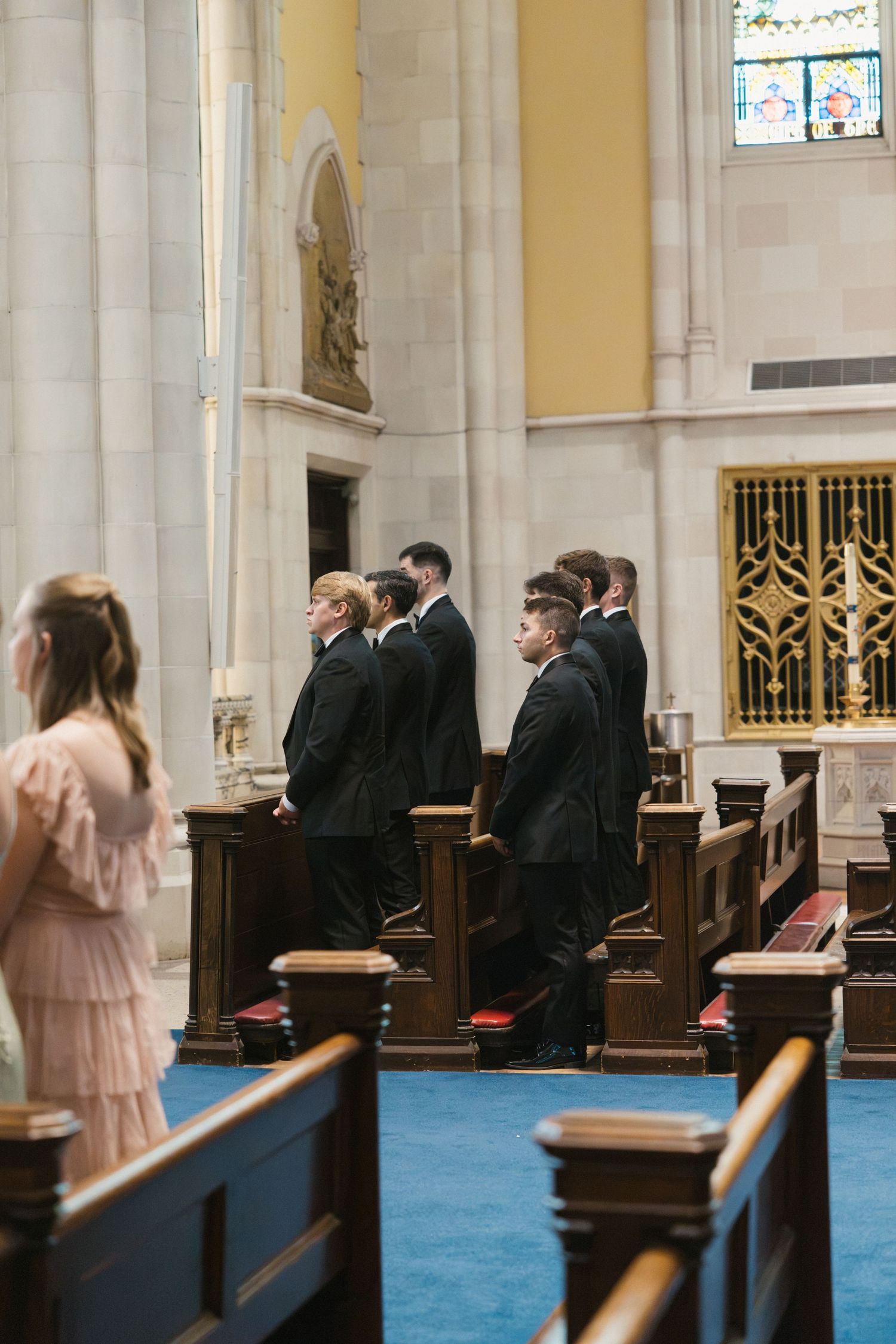Groomsmen in black suits standing in church pews during a wedding ceremony with stained glass windows.