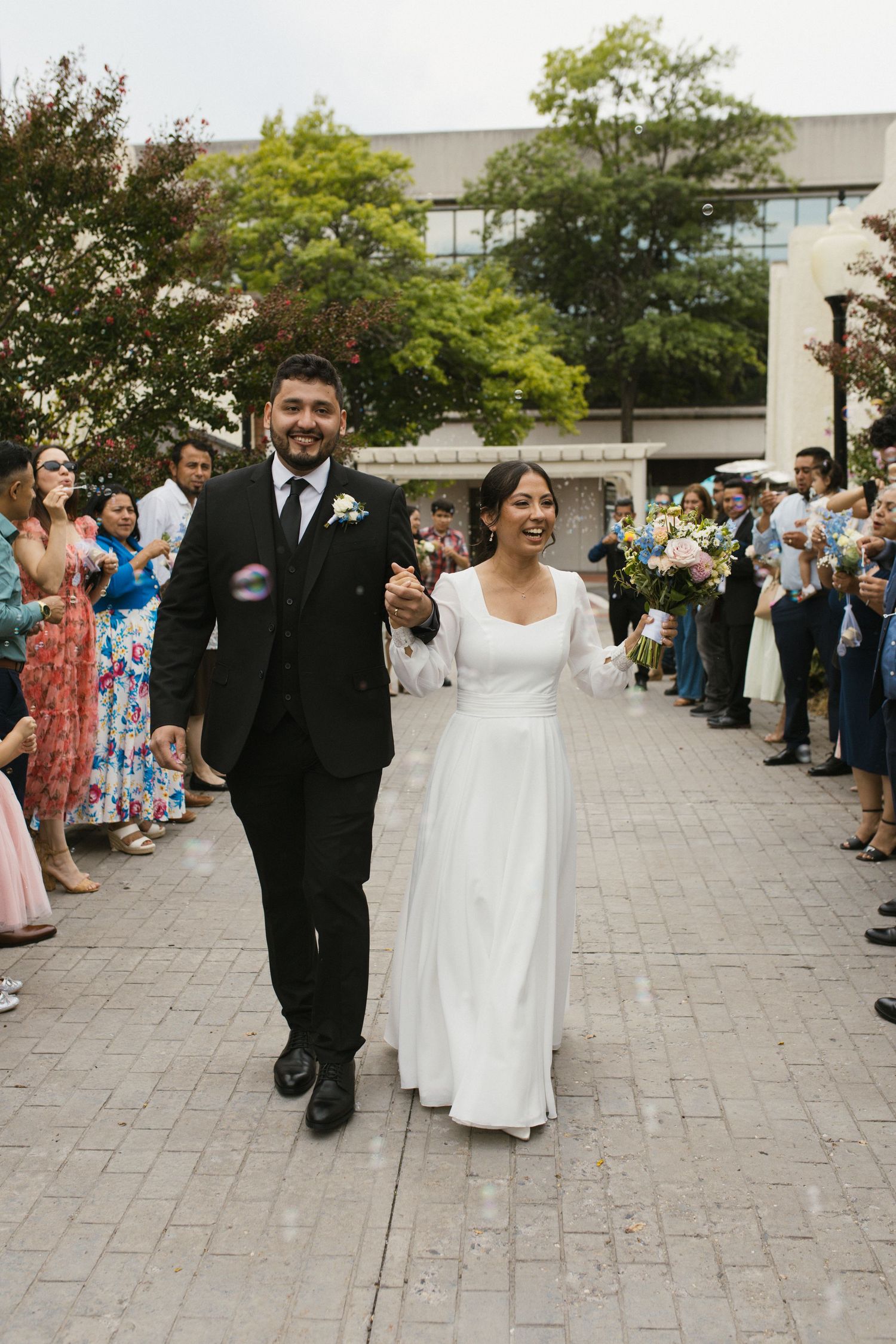 Smiling bride and groom exit church ceremony through joyful bubble sendoff crowd.