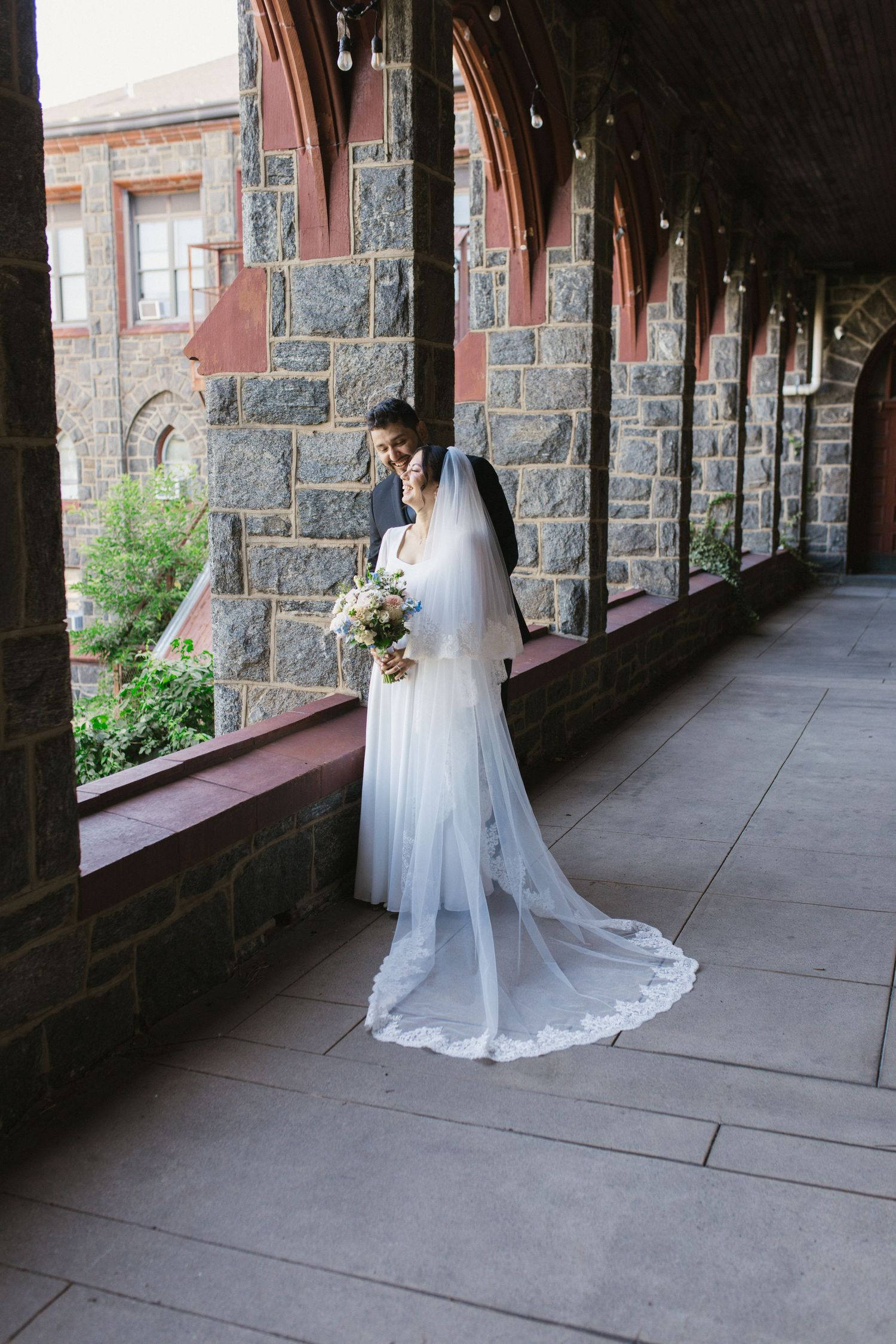 Bride in white gown and veil holds bouquet beside groom in stone archway corridor of historic building.
