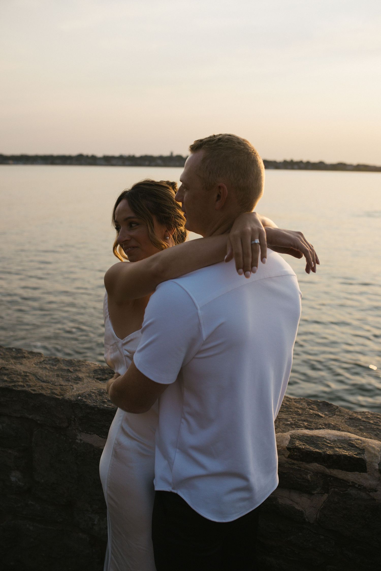 Couple sharing a romantic embrace on rocky waterfront at golden hour sunset.