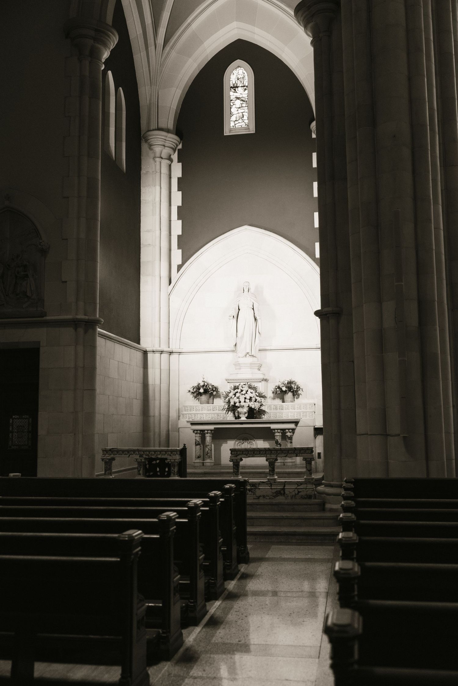 Black and white interior of a Catholic church with wooden pews leading to a statue at the altar.