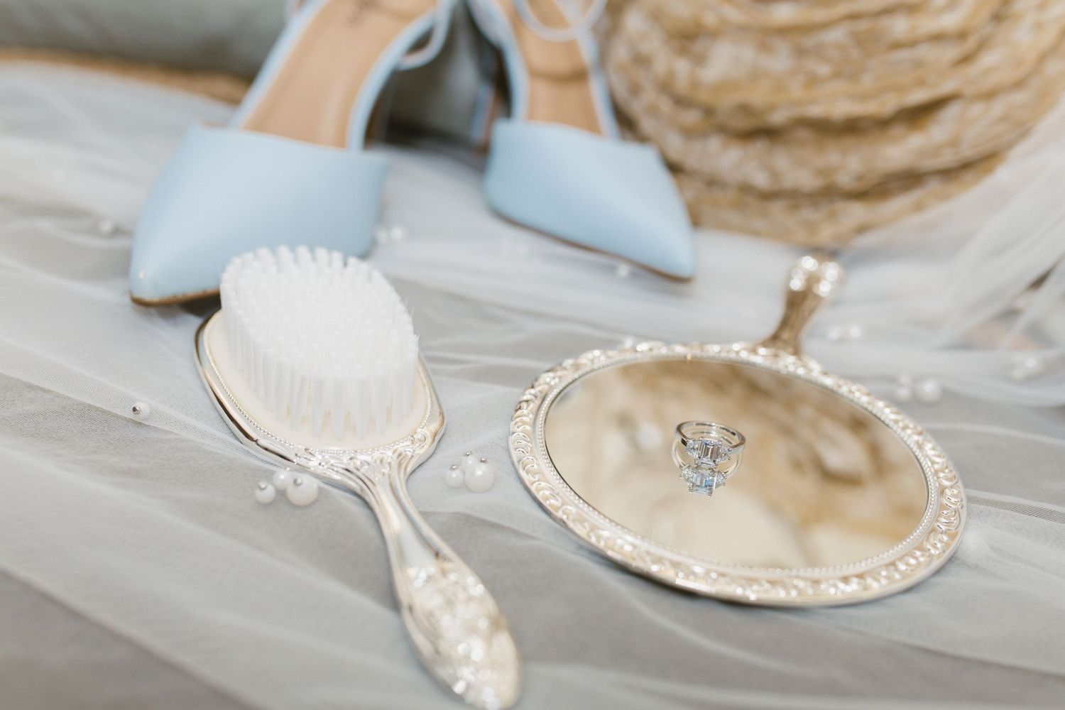 Silver vintage hand mirror and brush beside diamond ring and light blue bridal heels on veil. Catholic wedding.