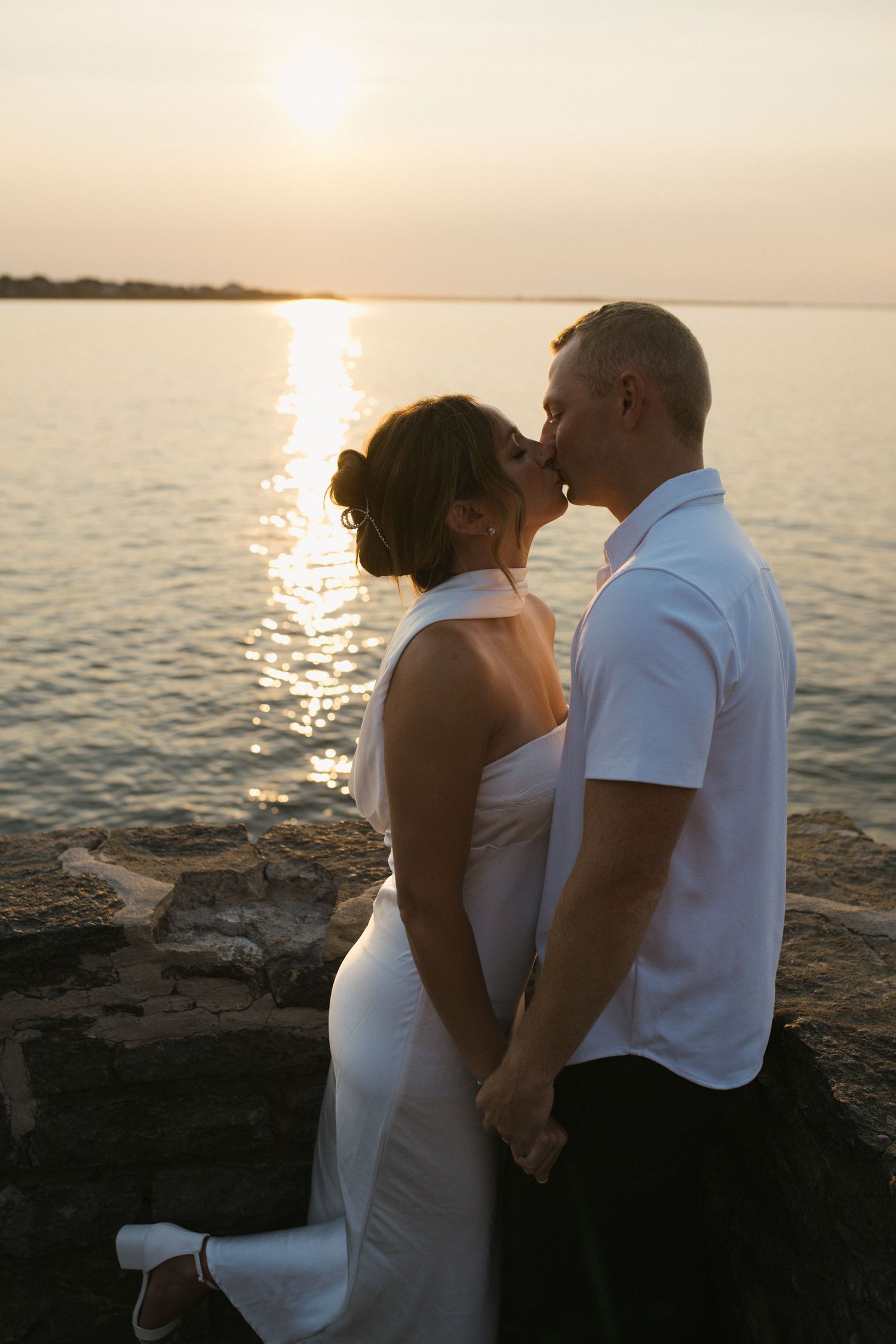 Couple sharing a kiss while holding hands on rocky shoreline during golden sunset.
