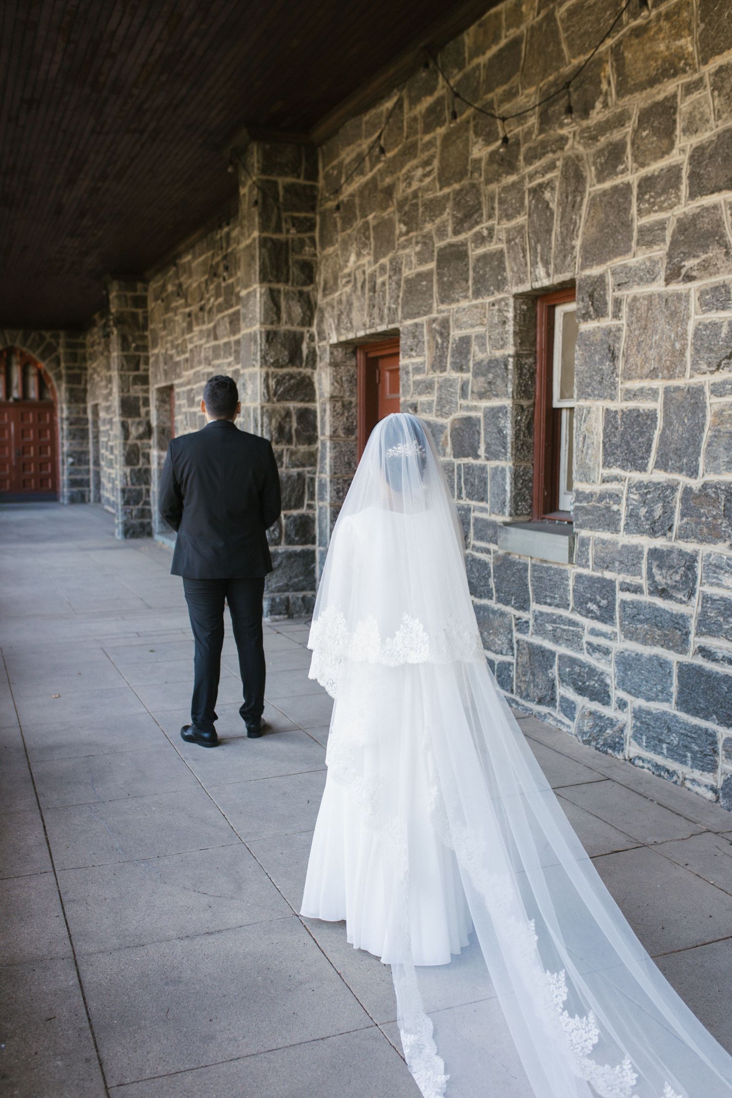 Bride in white gown and veil walks with groom in black suit along stone church corridor.
