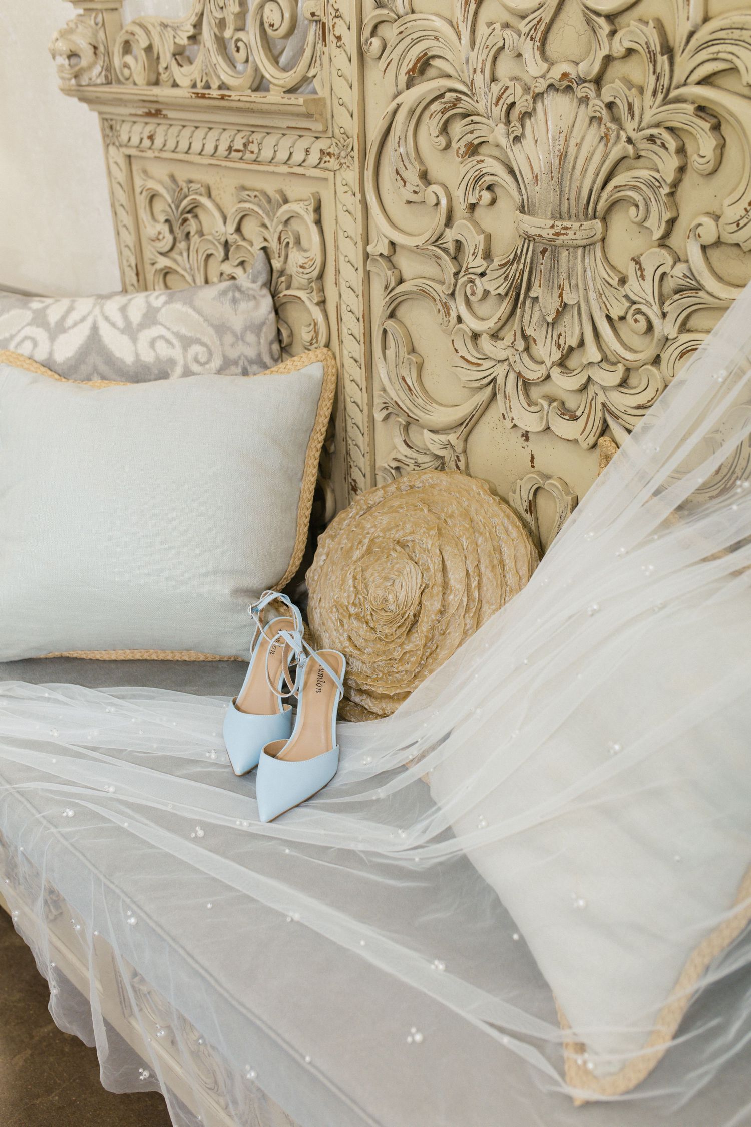 Light blue bridal heels rest on a tulle veil beside pillows on an ornate carved vintage settee.