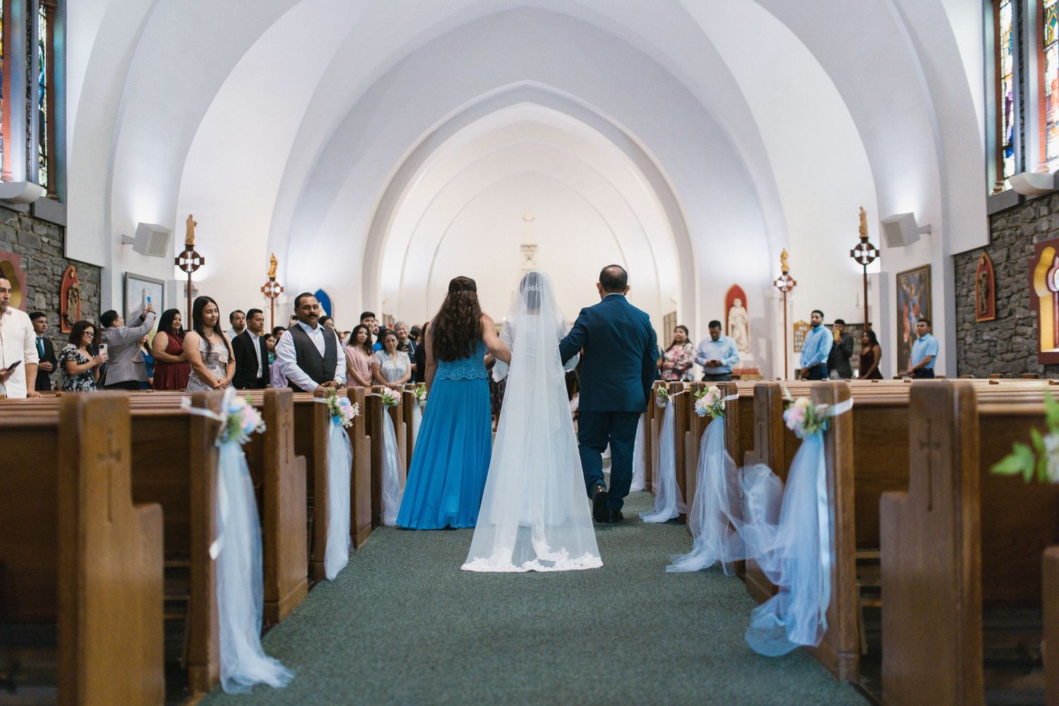 Bride walks down the aisle of a traditional church with wood pews, accompanied by wedding party in blue dresses.