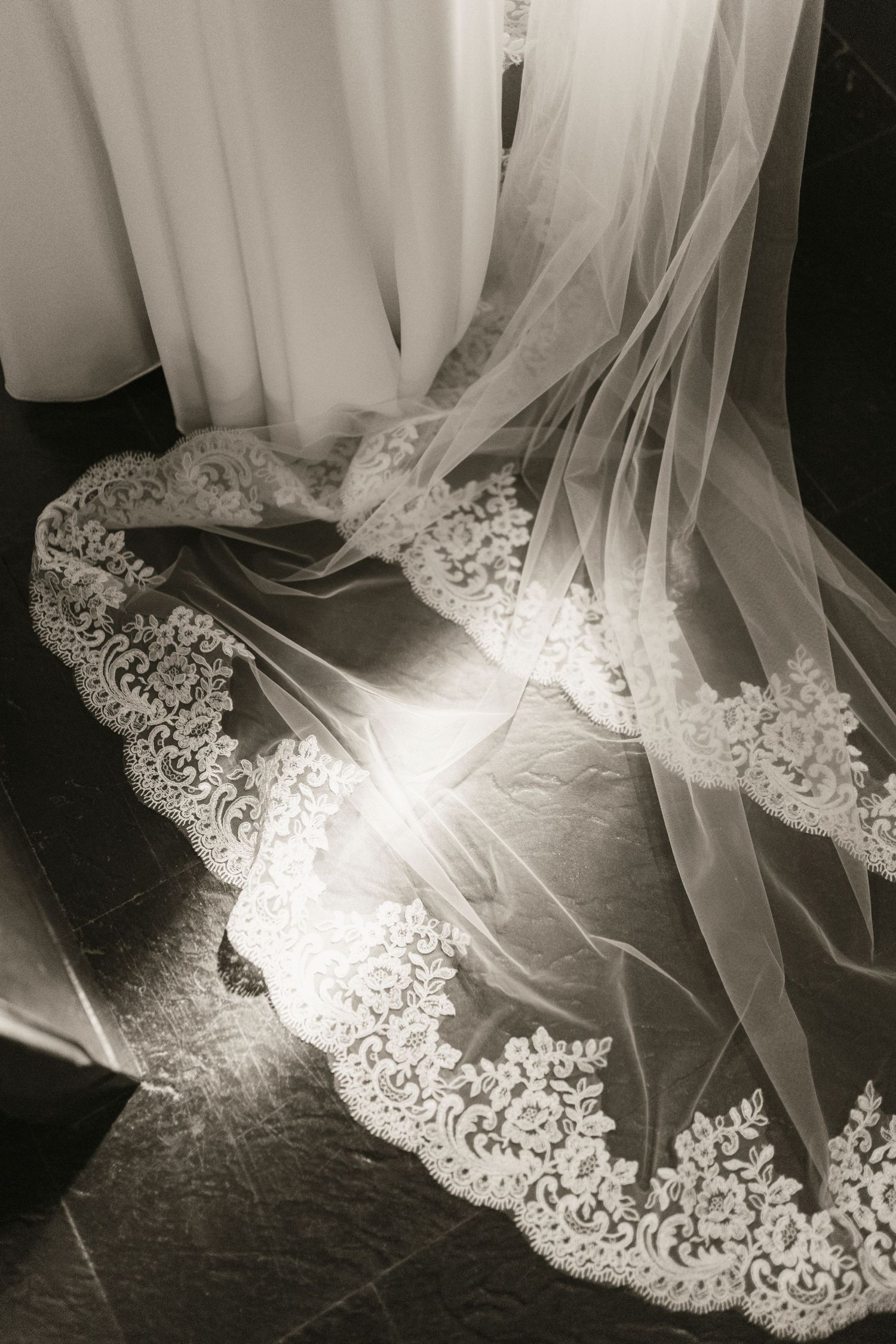 Black and white close-up of elegant lace-trimmed bridal veil with soft light on dark floor.