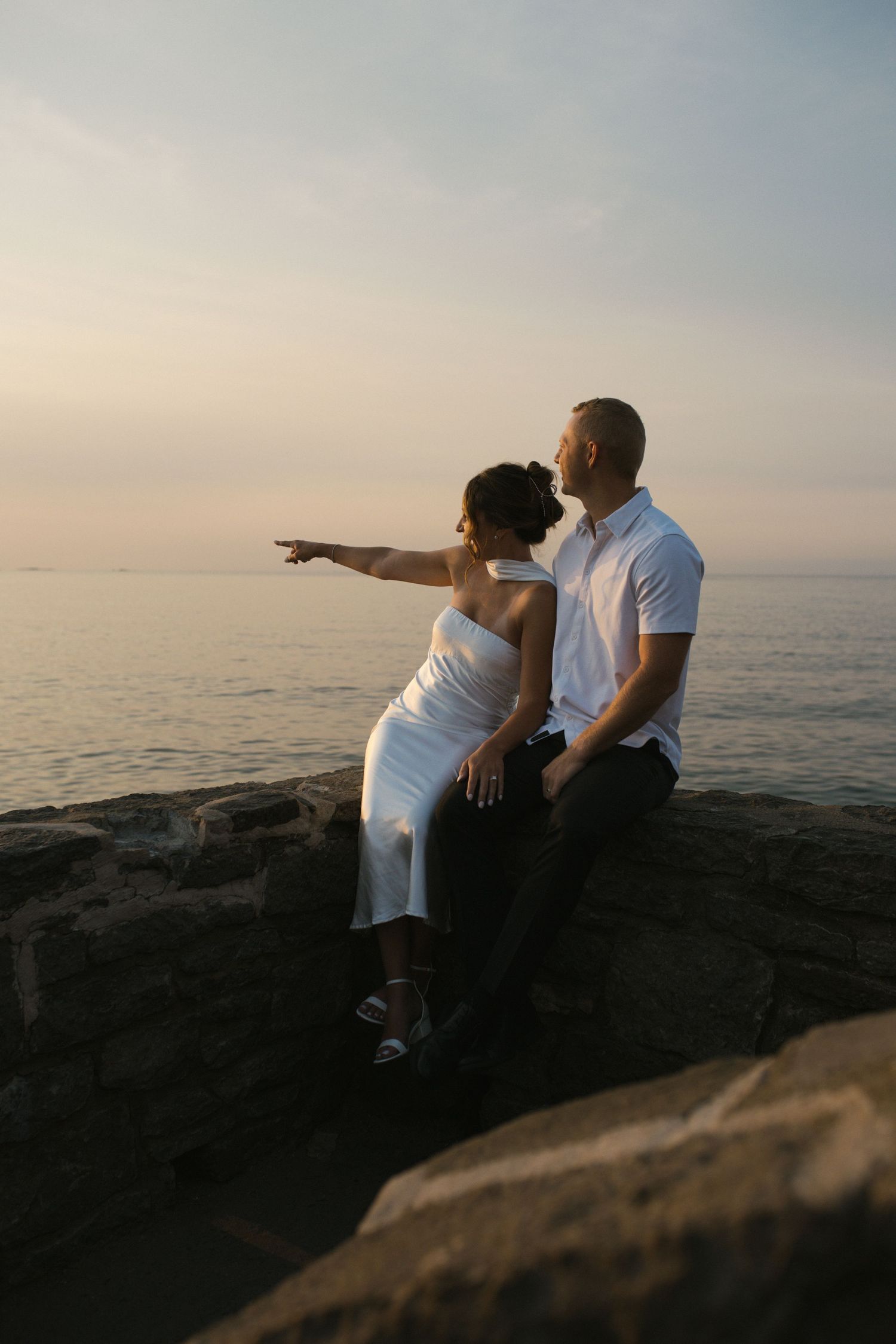Couple sitting on rocky seaside wall at sunset, woman pointing toward the horizon over calm ocean.