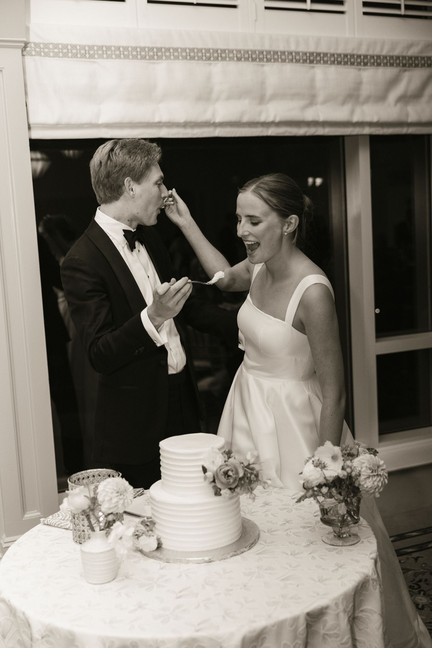 Bride feeds groom wedding cake while laughing beside white tiered cake and floral arrangements at reception.