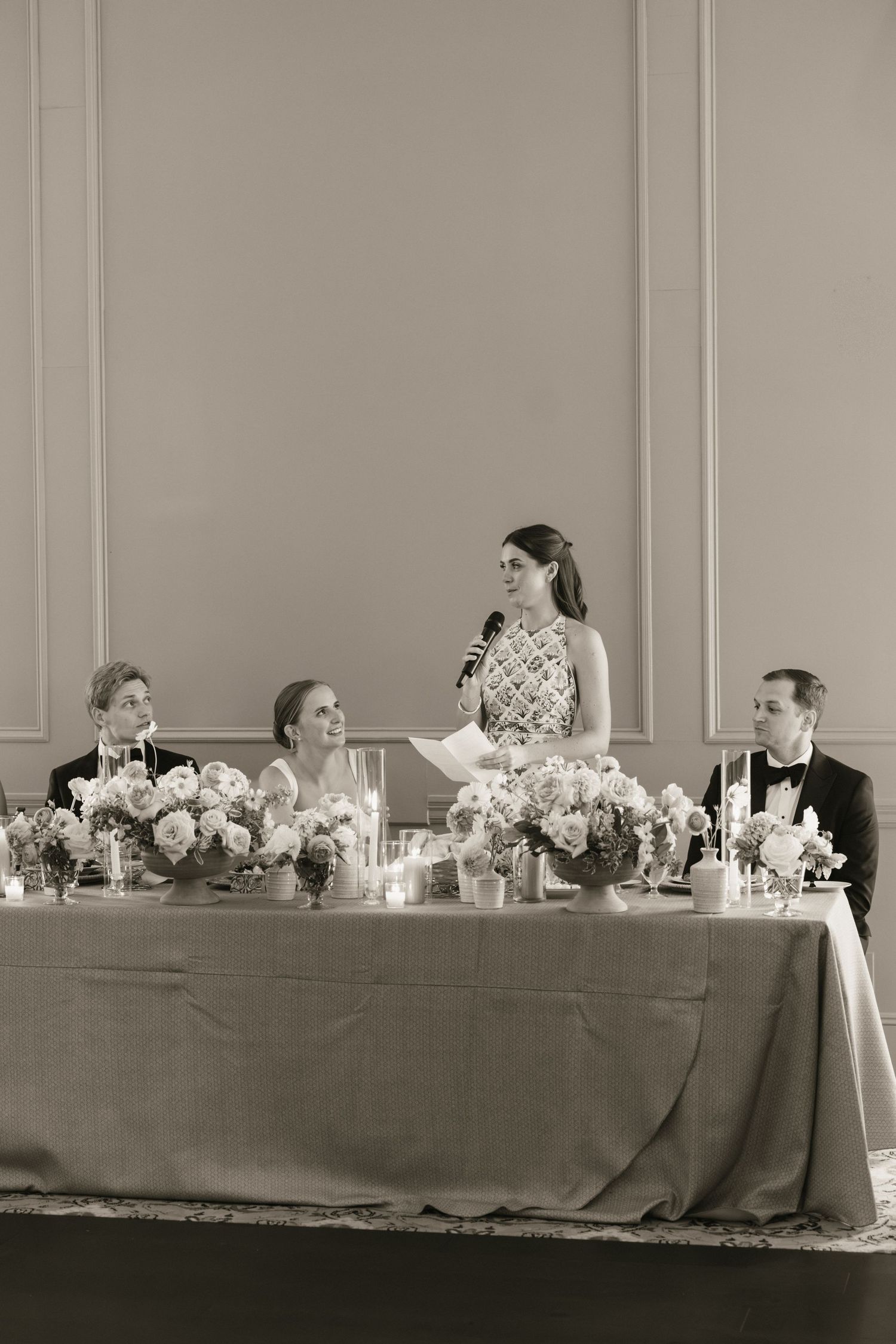 Black and white photo of bridesmaid delivering speech at wedding head table with floral arrangements.