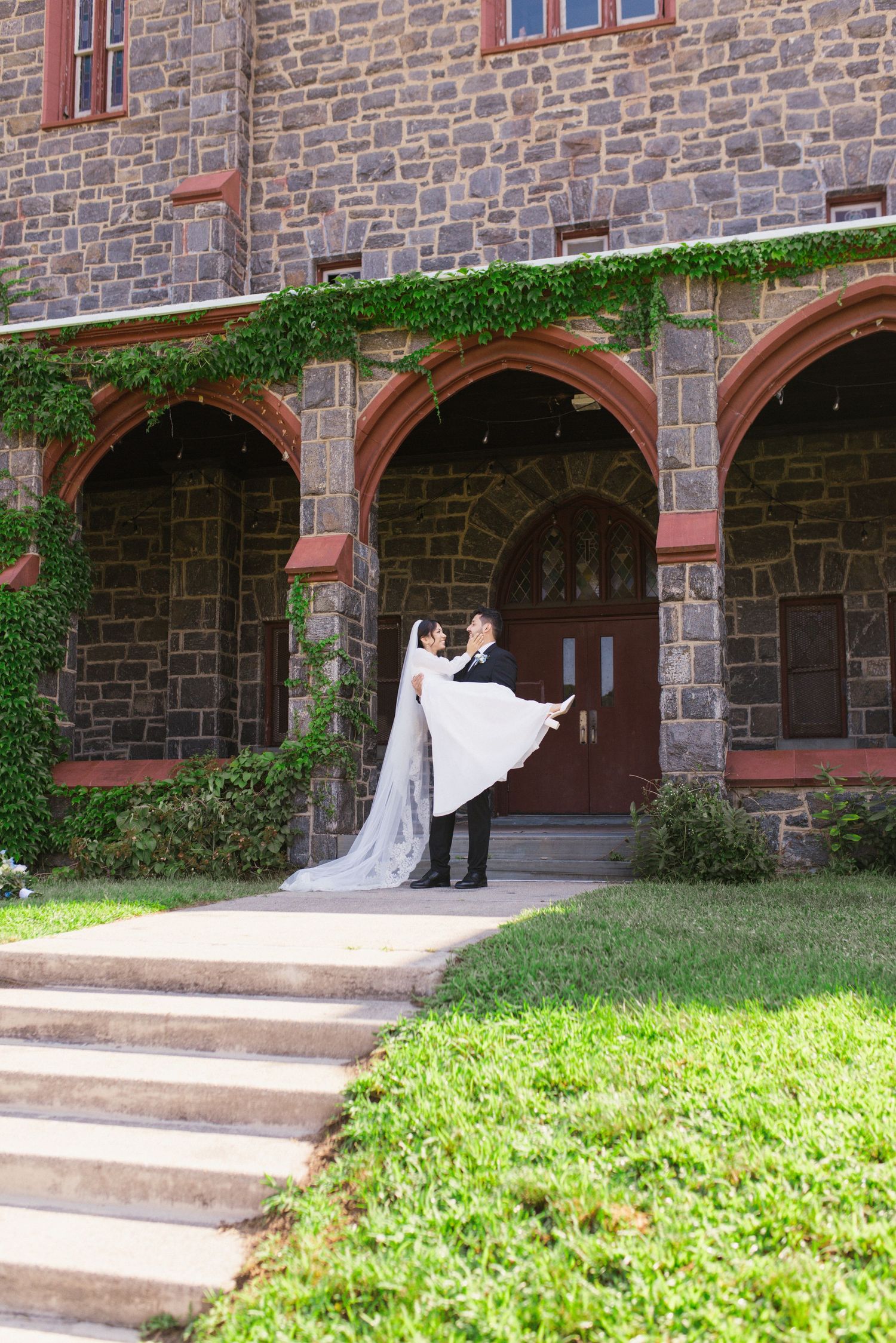 Groom lifting bride in wedding dress outside stone church with ivy-covered arches and red wooden doors.