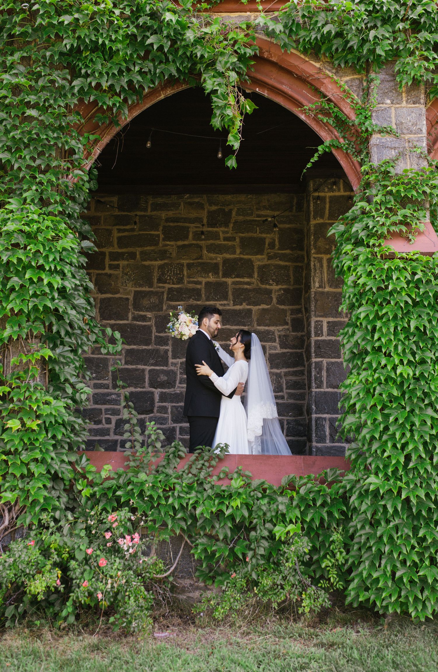 Bride and groom embracing in an ivy-covered stone archway during their outdoor wedding ceremony.
