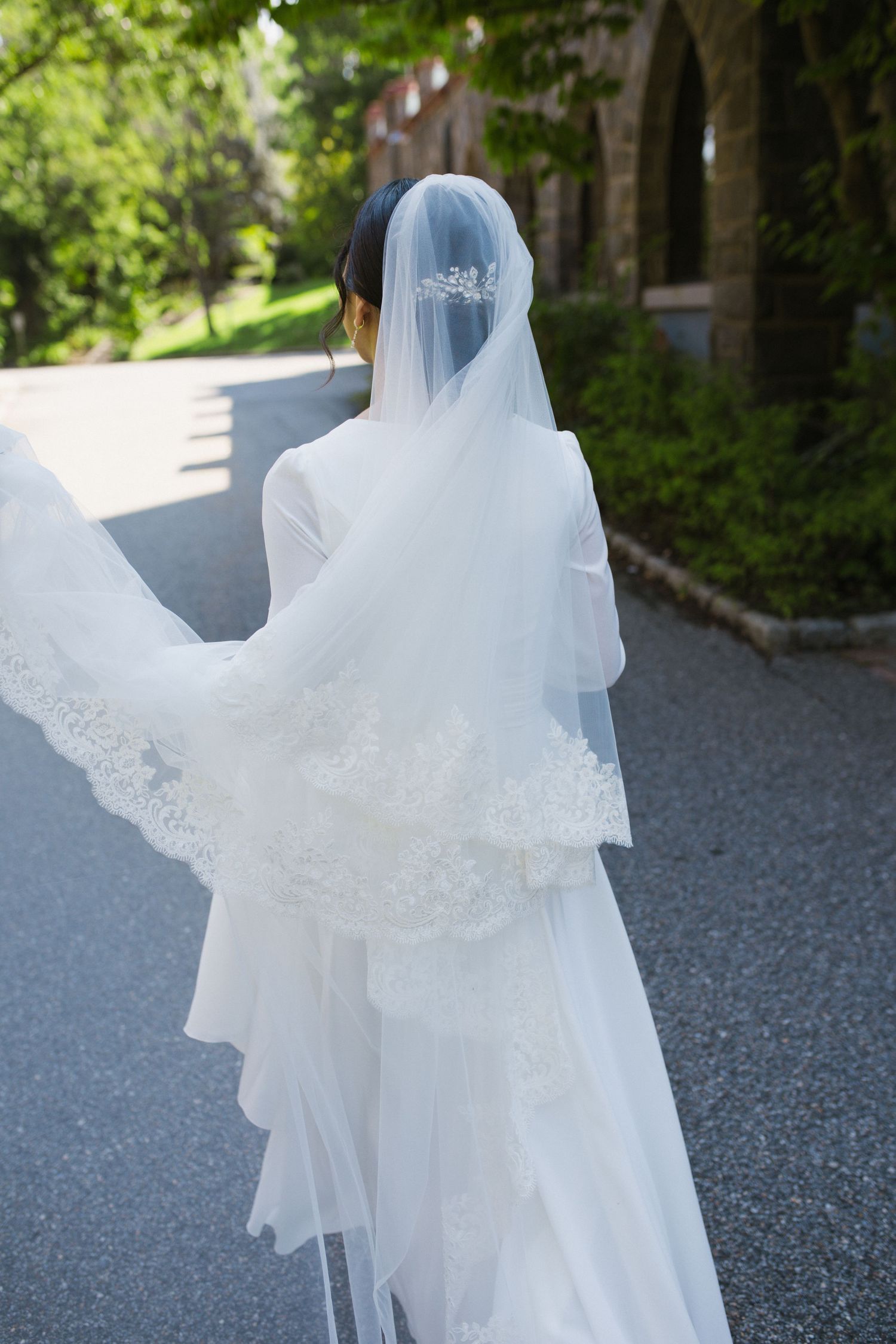 Bride walking in a flowing white wedding gown with lace-trimmed cathedral veil outdoors.