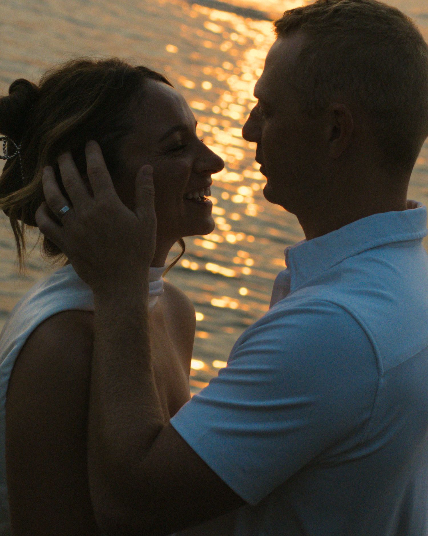Couple sharing a romantic moment at sunset by the water, woman wearing engagement ring, golden light reflecting behind them.