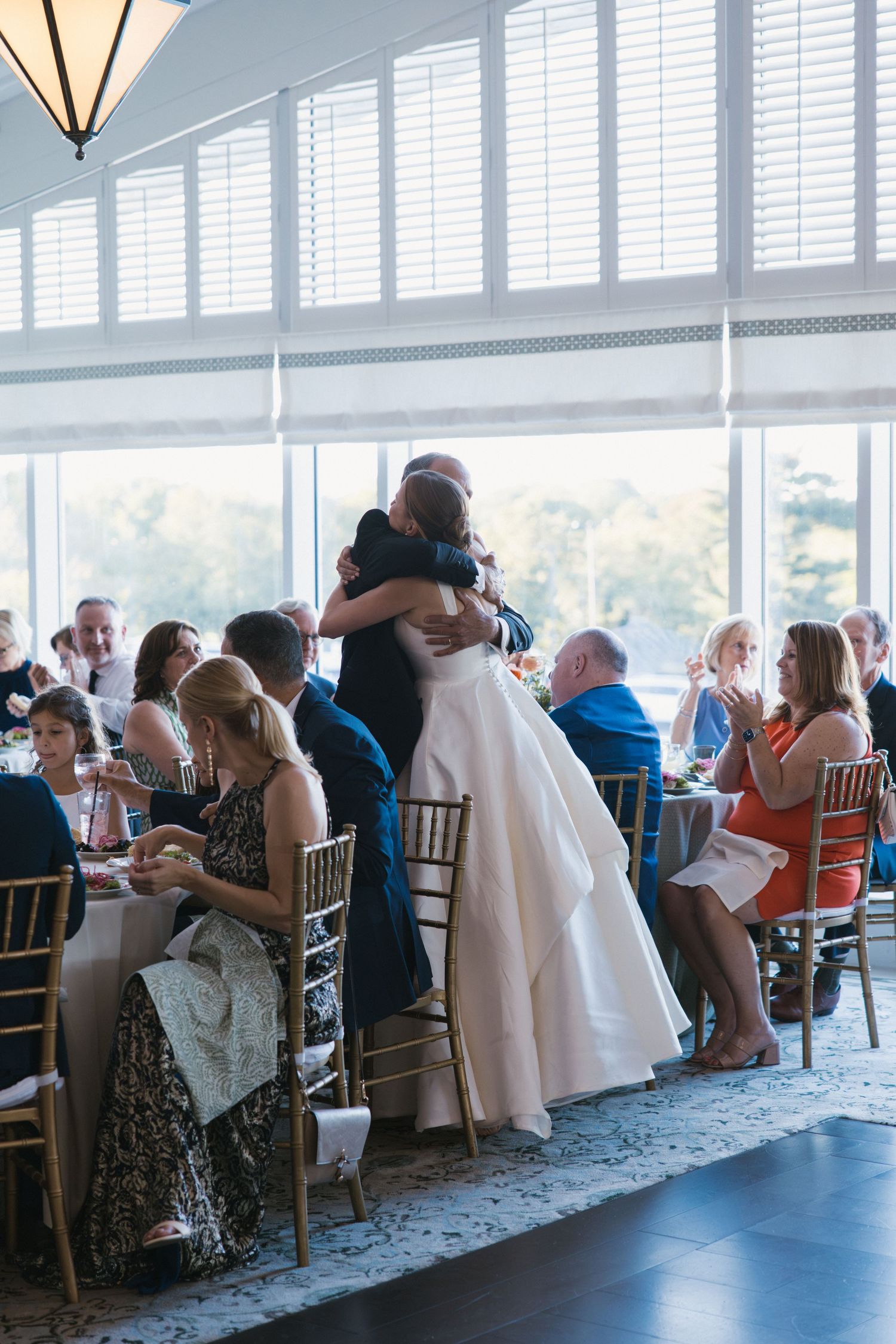 Bride in white ball gown embraces a guest during wedding reception in a bright windowed venue.