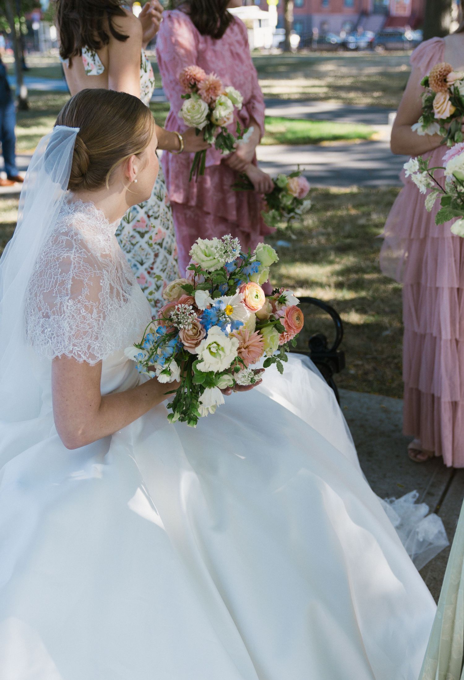 Bride in white lace veil holds colorful floral bouquet while seated outdoors with bridesmaids nearby.