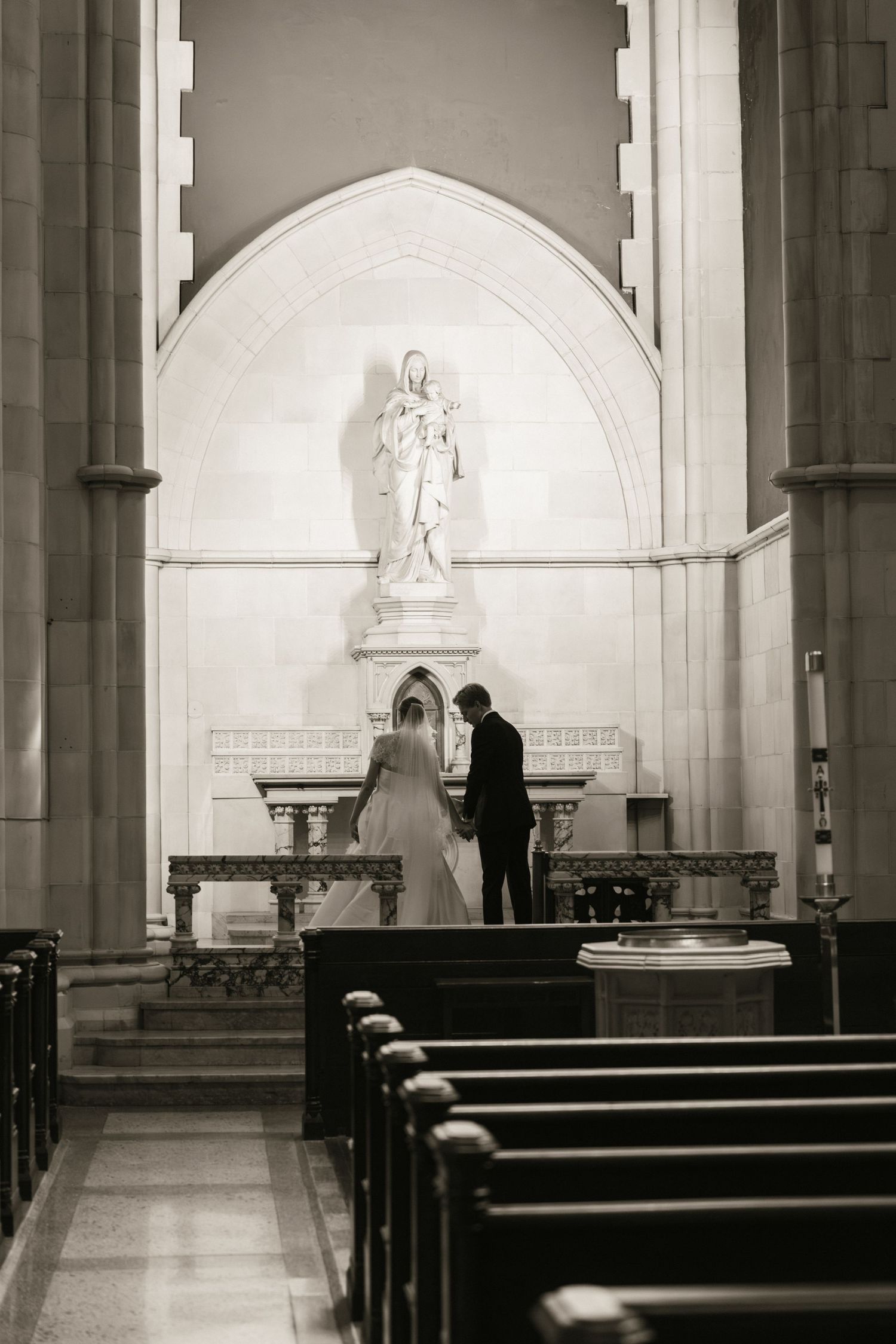 Black and white photo of bride and groom standing before a statue in a church side chapel.