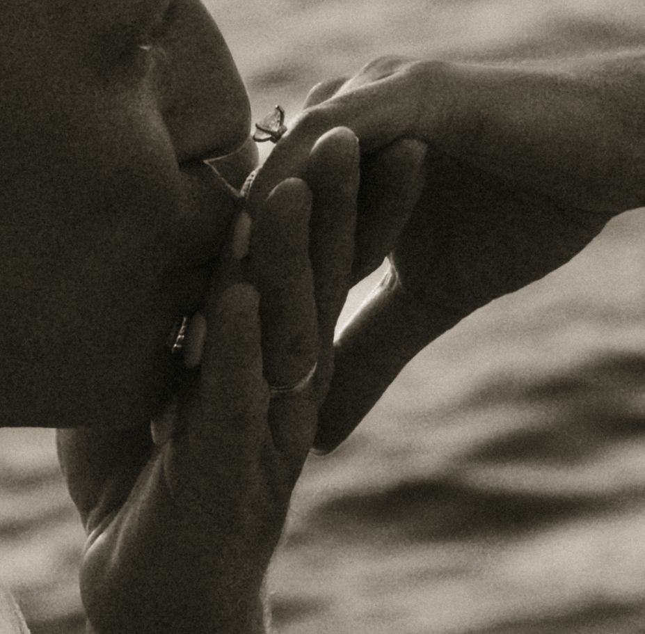 Close-up sepia photo of two hands tenderly touching near a face, with water in the background.