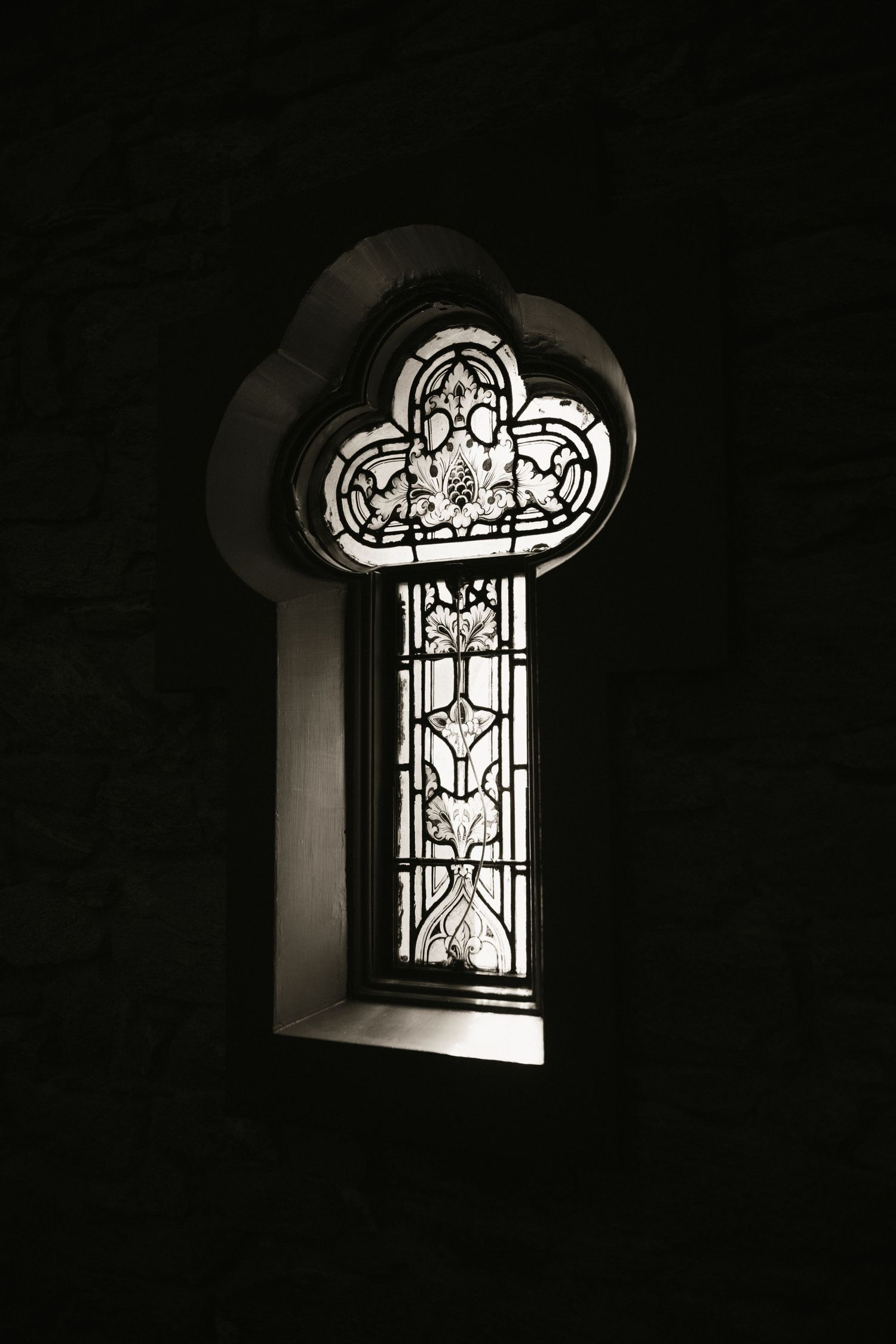 Black and white photo of ornate stained glass window in a dark stone church wall.