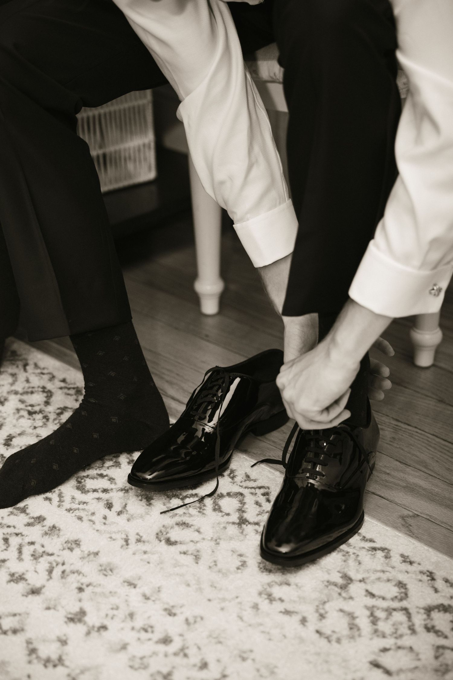 Groom tying black patent leather dress shoes on patterned rug during wedding preparations.