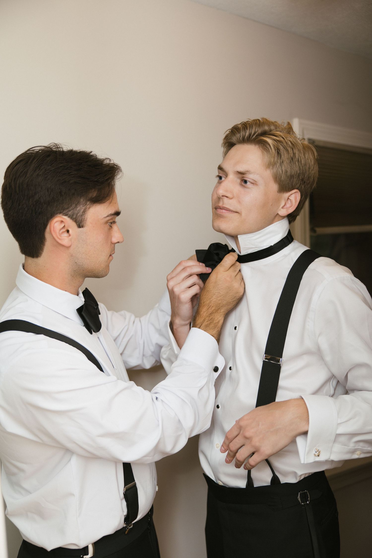 Two groomsmen in white shirts and black suspenders help each other with bow ties while getting ready for a wedding.