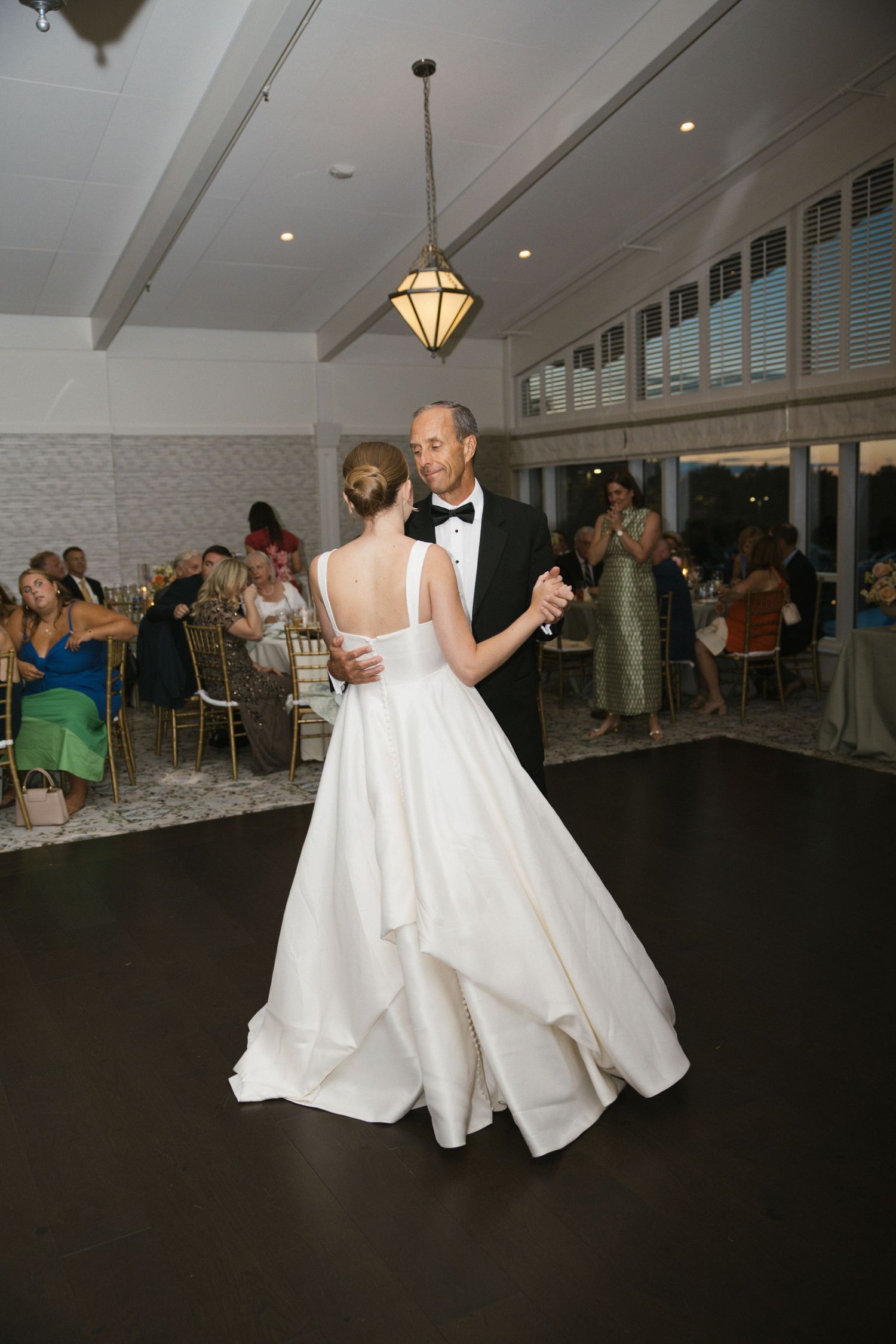 Bride and groom share their first dance in a bright reception hall while guests watch and celebrate in the background.