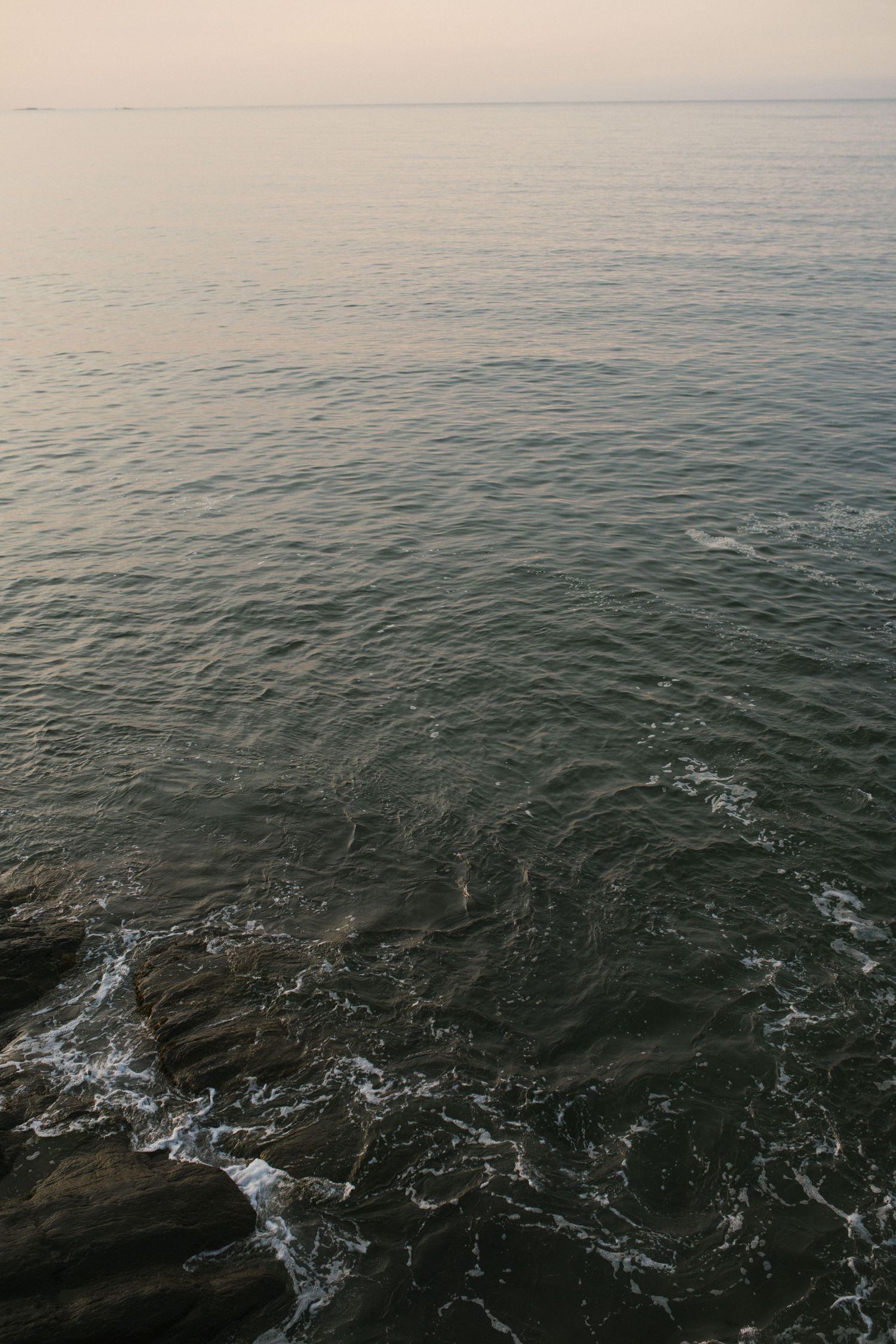 Rocky shoreline meets calm ocean waters at dusk, with gentle waves and a hazy horizon in this serene seascape.