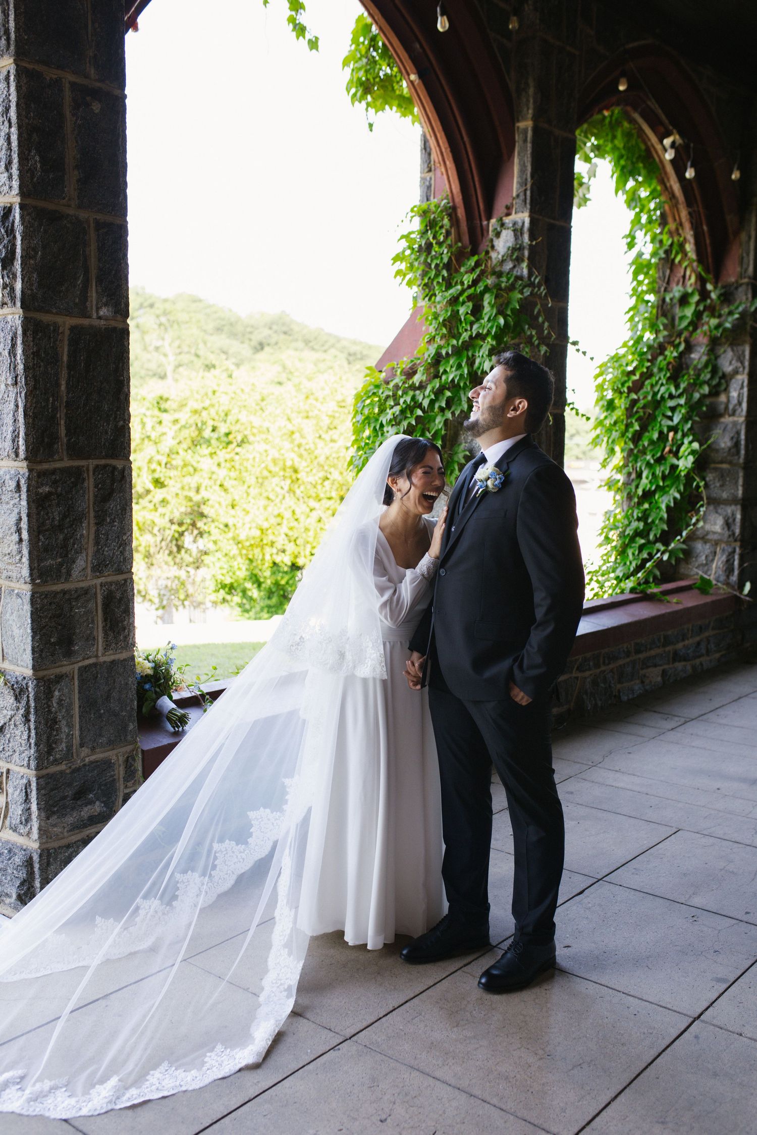 Bride and groom pose together in ivy-covered stone arch with lush green trees behind.