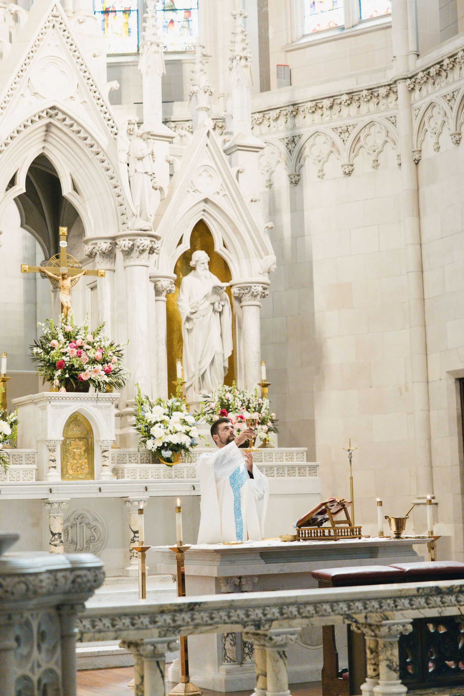 Priest performing wedding ceremony at ornate white Gothic cathedral altar with floral arrangements.