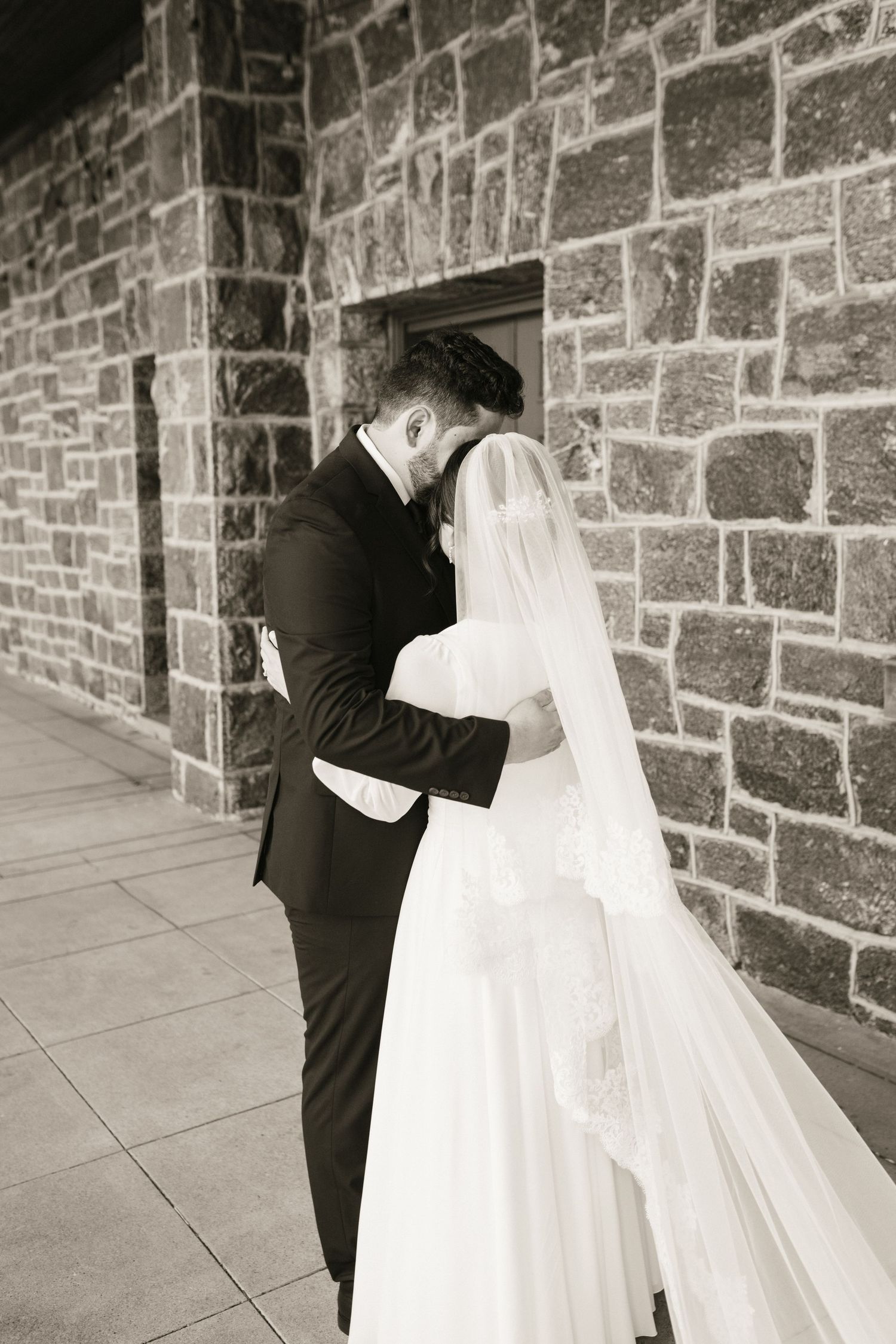 Black and white photo of bride and groom embracing against a stone wall, wedding veil flowing.