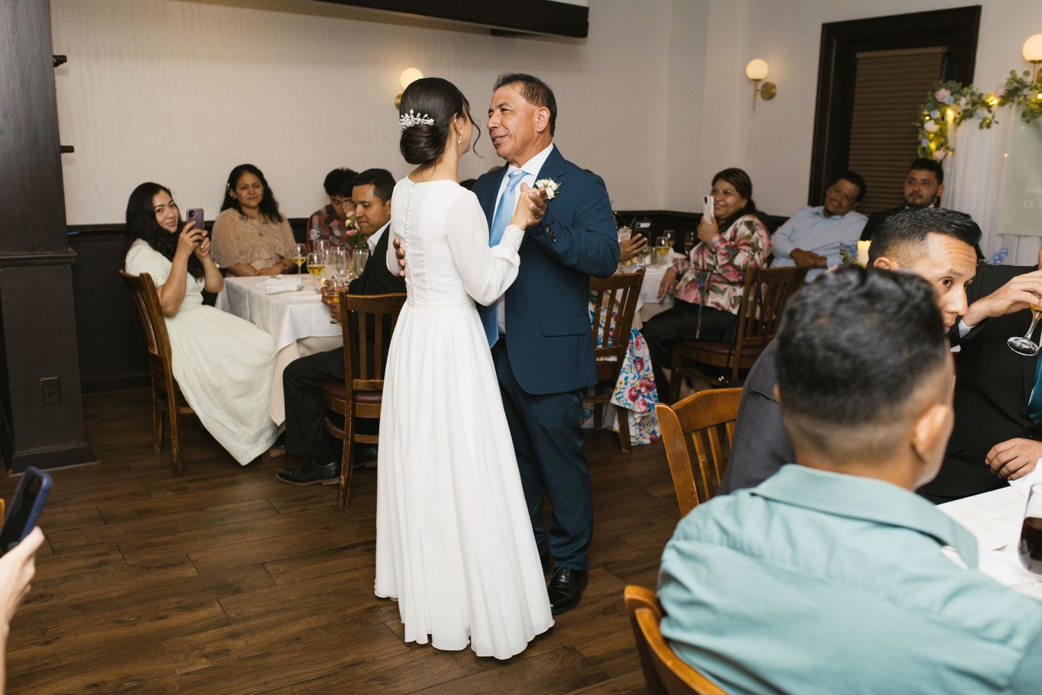 Bride shares a father-daughter dance at intimate wedding reception with seated guests.