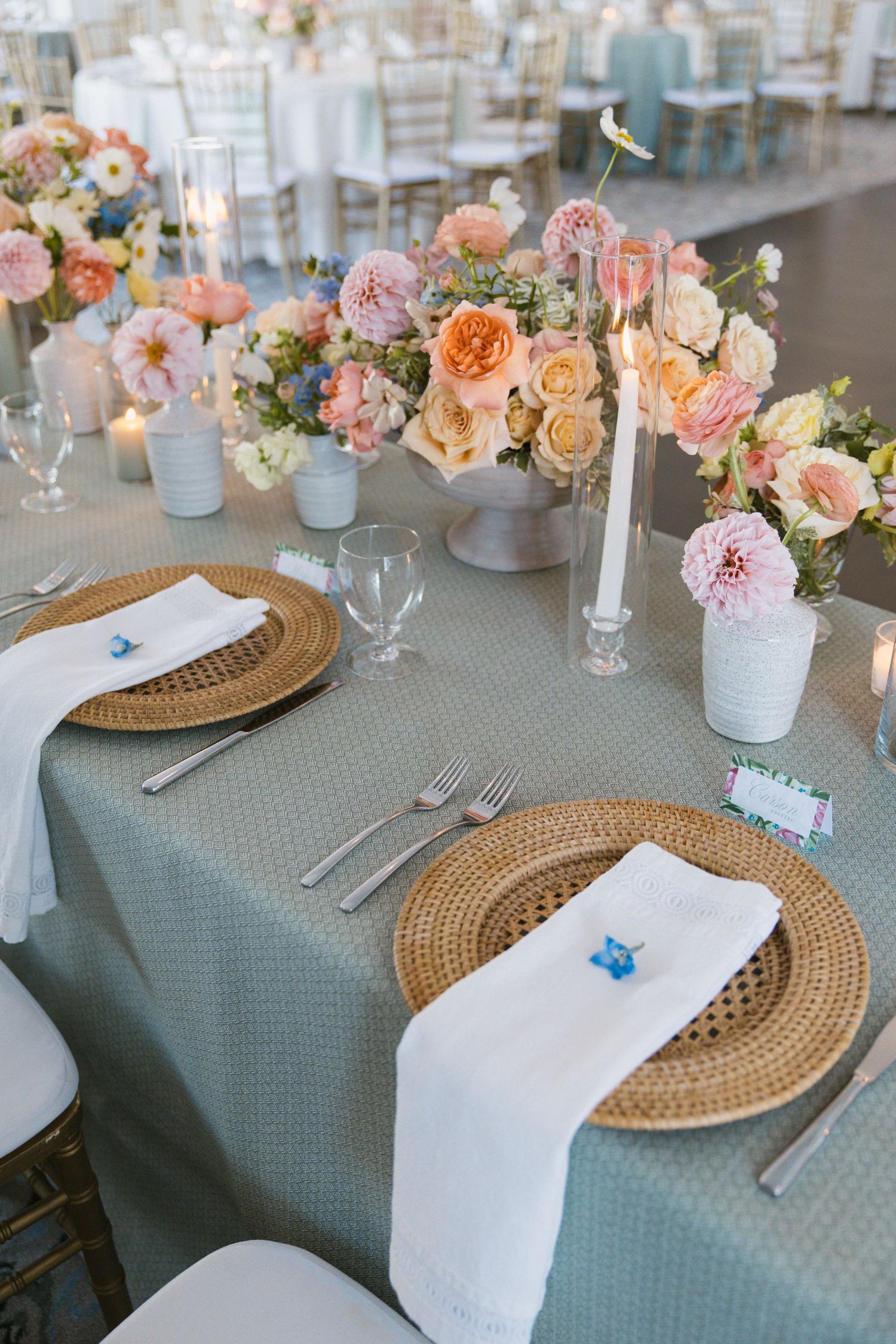 Elegant wedding reception table with woven charger plates, white napkins, and lush pink and peach floral centerpieces on sage linen.