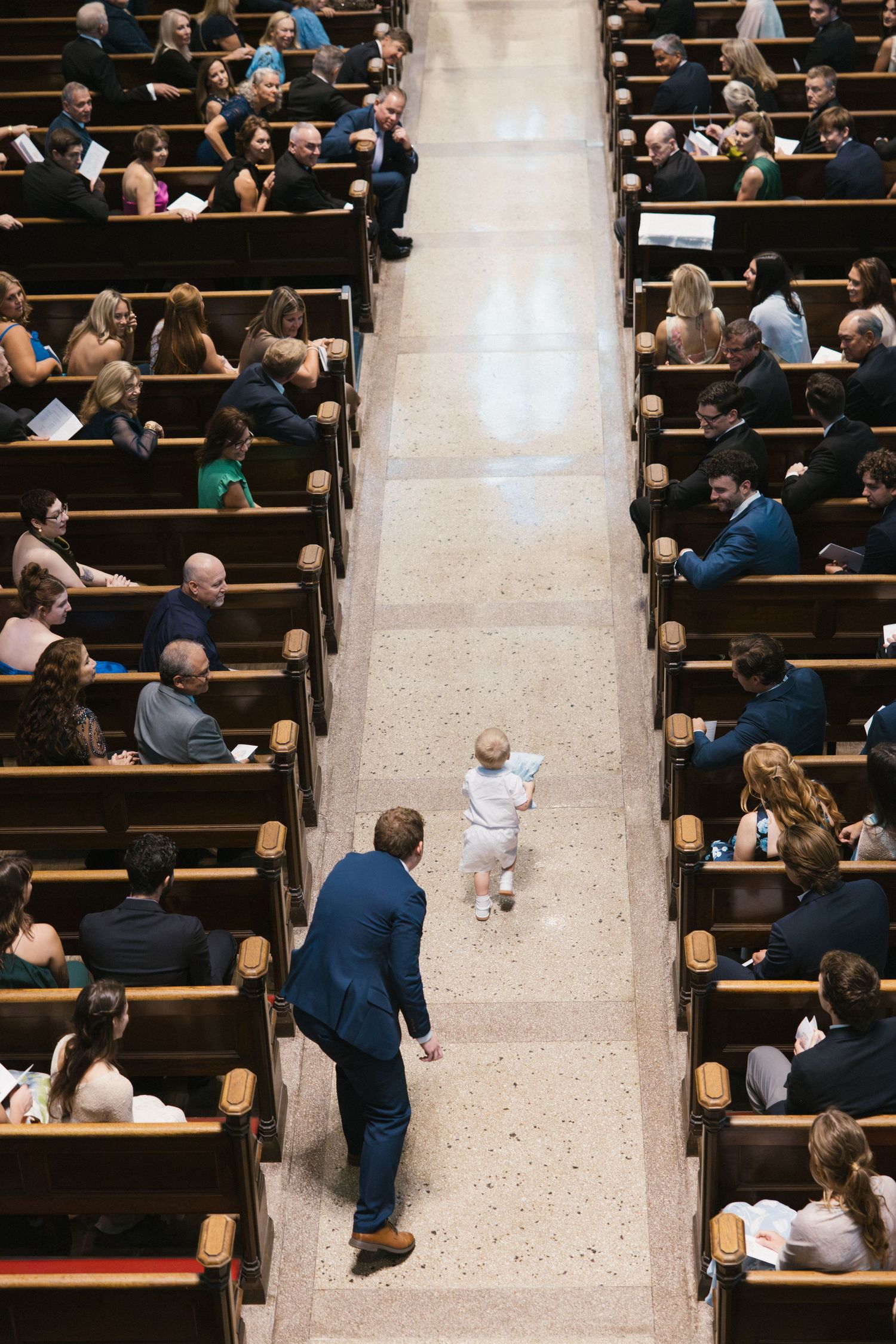 A toddler walks down a church aisle during a ceremony while an adult in a blue suit watches nearby.