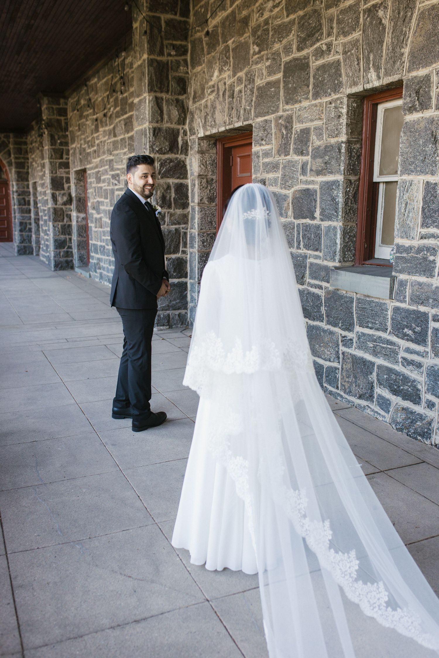 Bride in lace-trimmed cathedral veil stands before groom in black suit outside stone church building.
