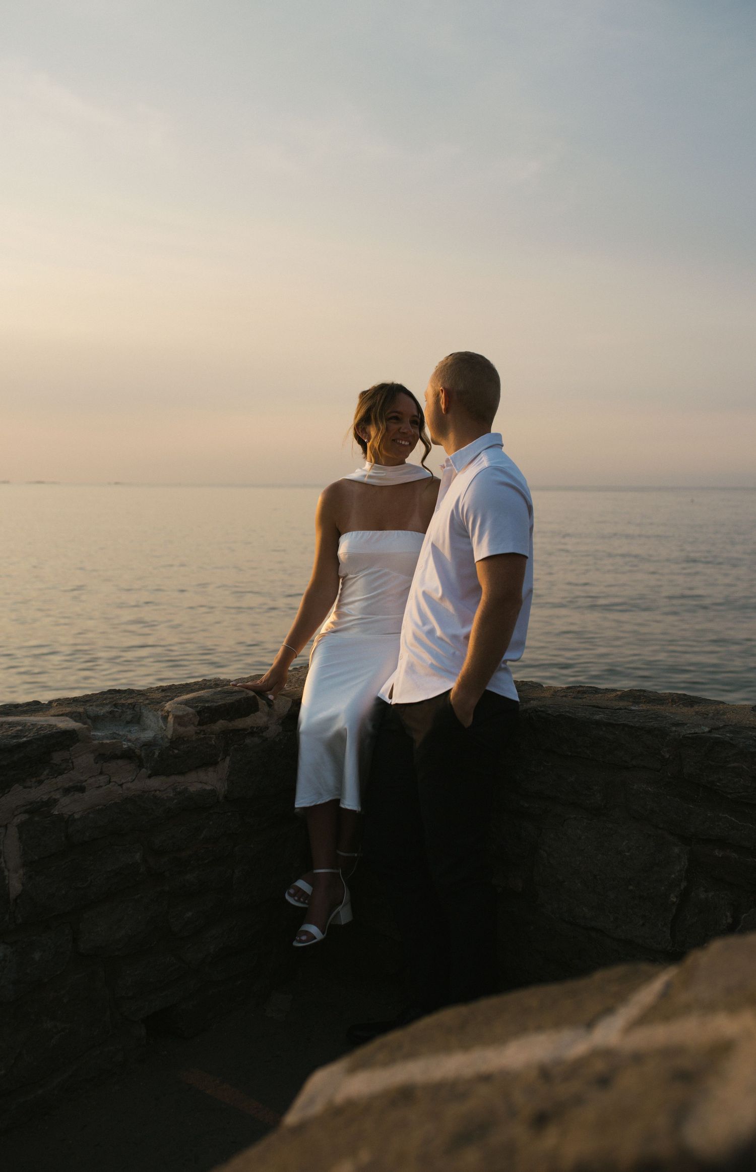 Couple posing on stone wall at sunset overlooking serene ocean, woman in white dress.