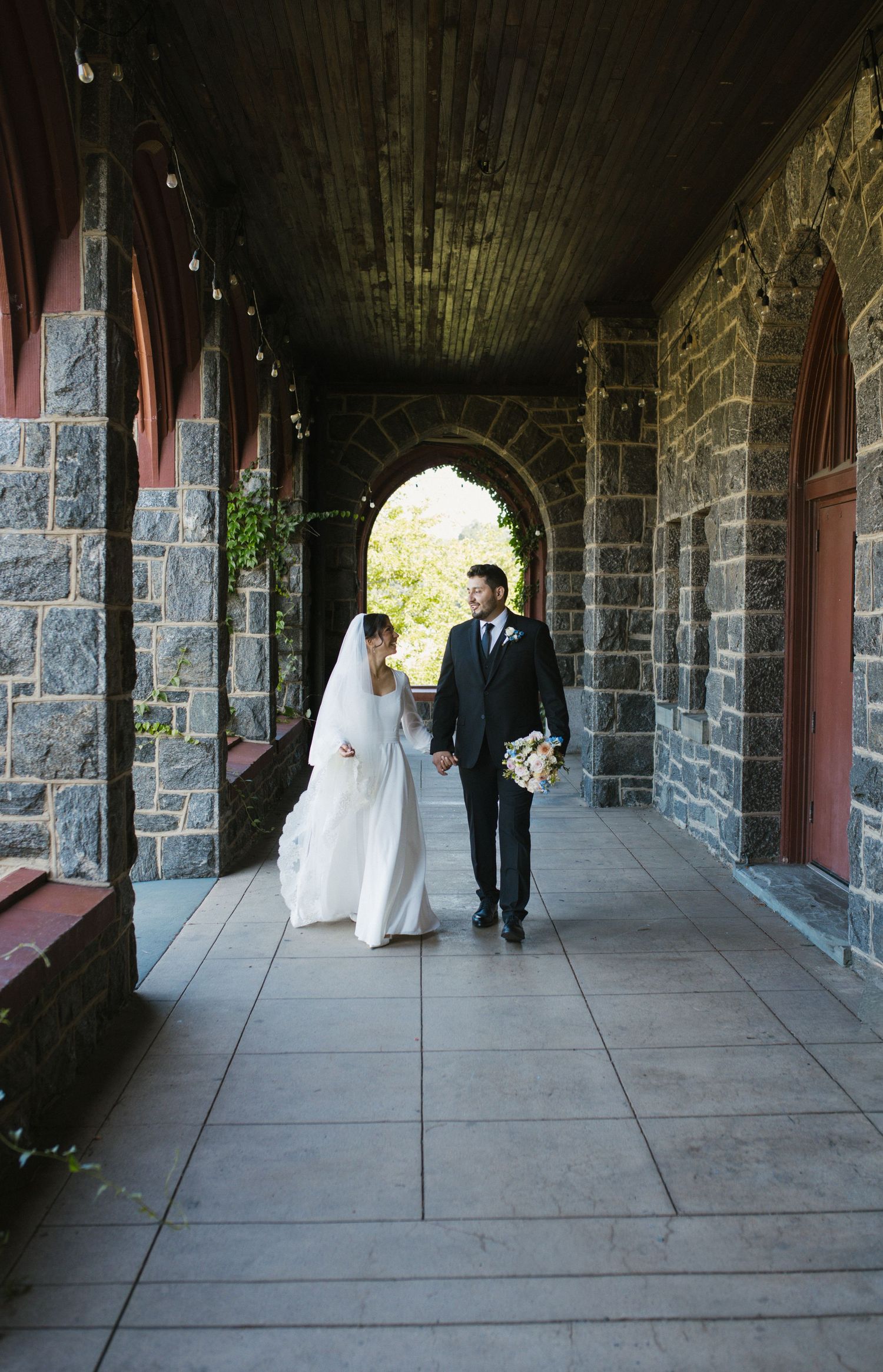 Bride and groom walk hand-in-hand through a stone archway corridor at their wedding venue.