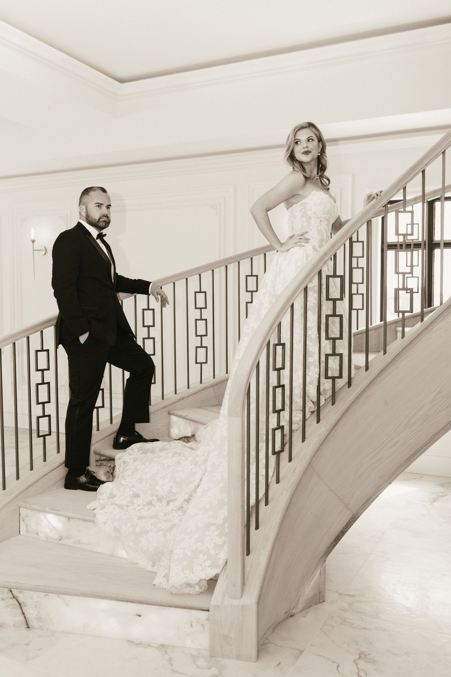 Black and white photo of bride and groom posing on elegant curved staircase in classic wedding attire.