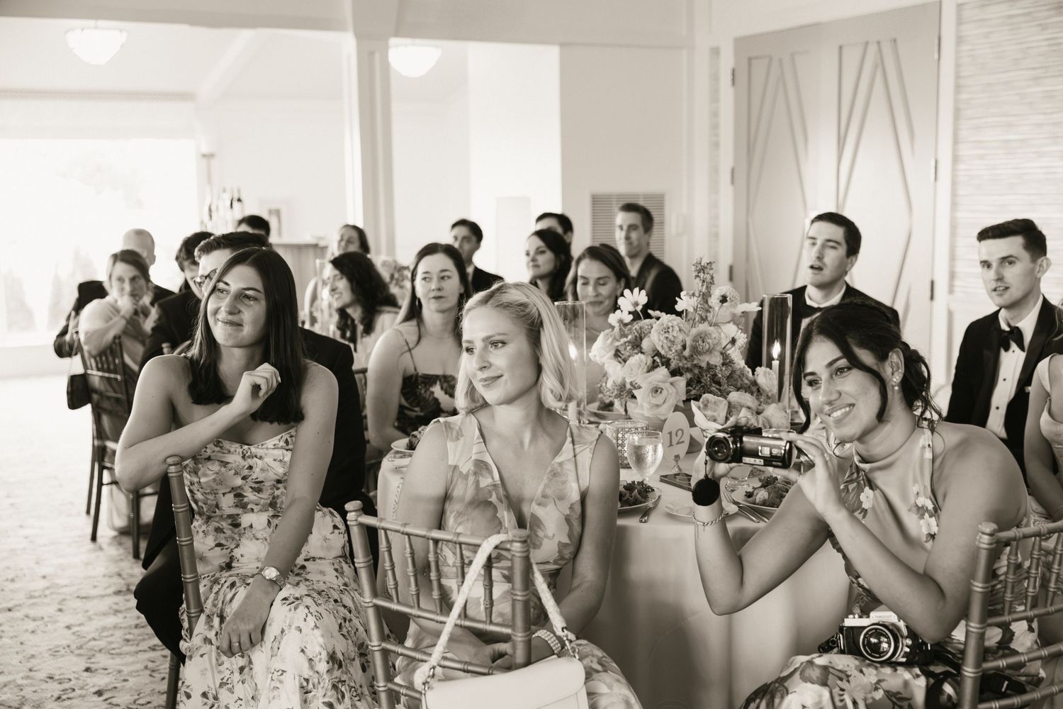 Wedding guests laughing and toasting during reception speeches in an elegant venue, captured in black and white.