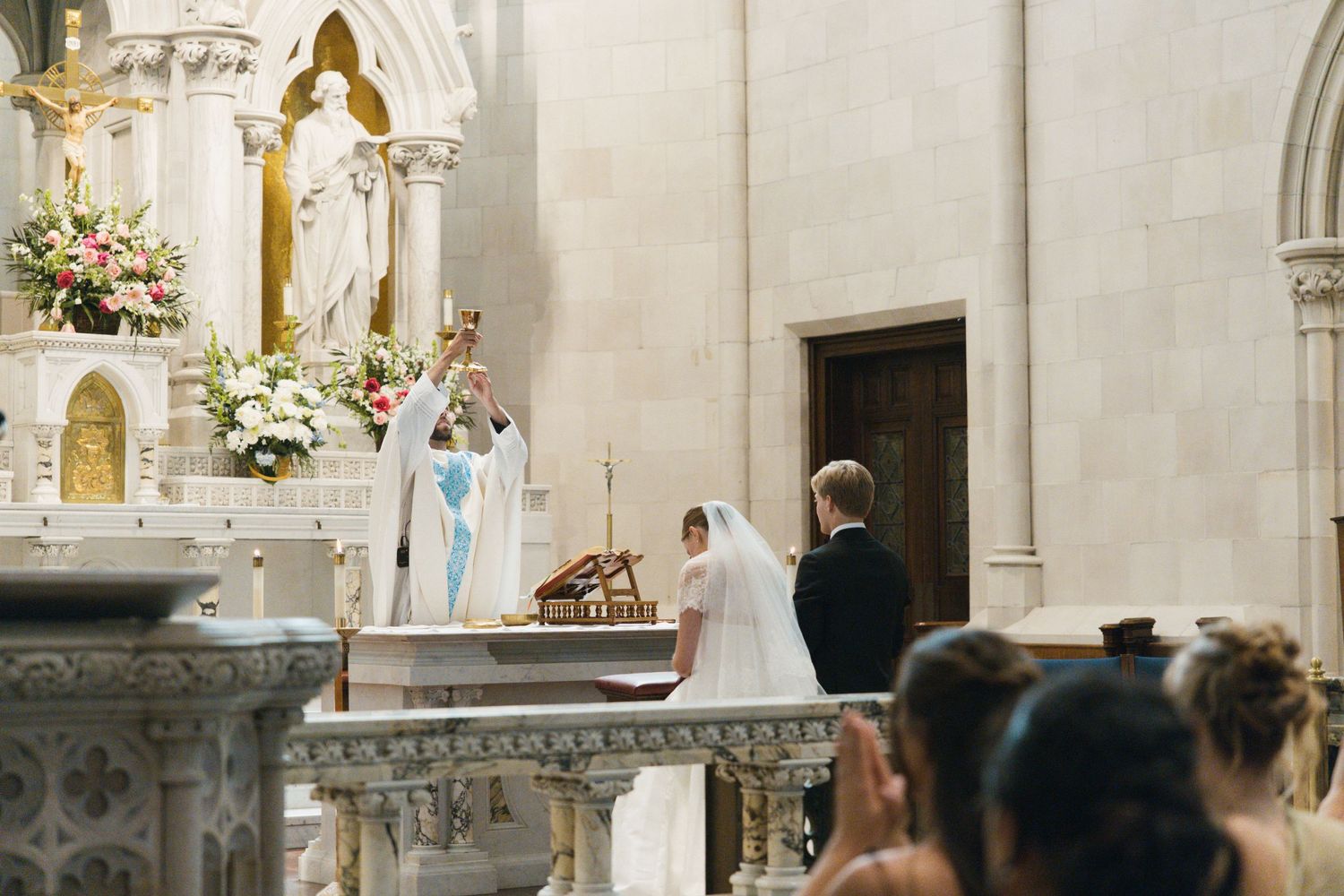 Catholic wedding ceremony with priest in blue vestments blessing bride and groom at ornate white marble altar.