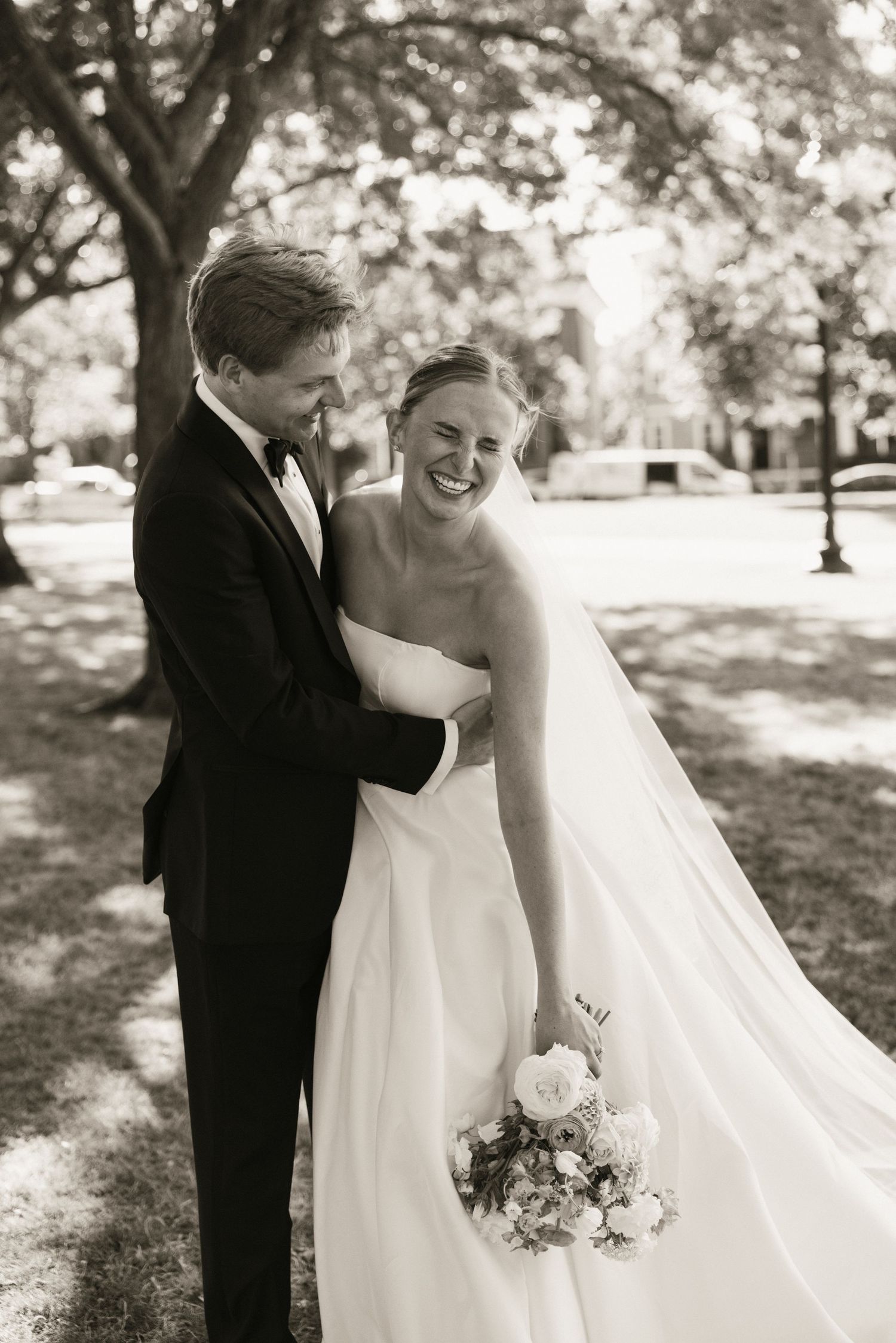 Black and white portrait of laughing bride holding bouquet embraced by groom in tuxedo outdoors.